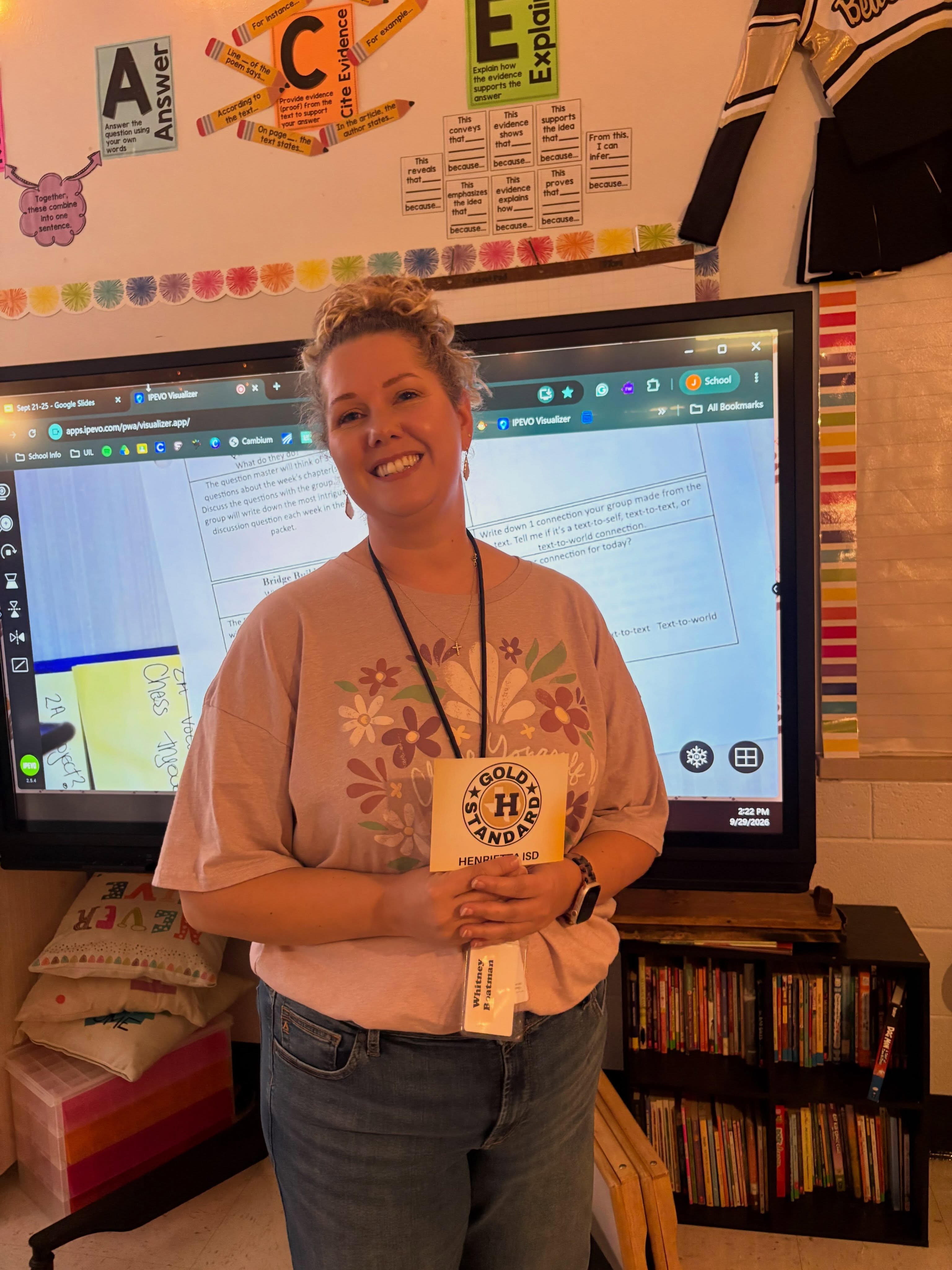 Woman standing in front of a classroom screen, smiling, with a name tag and a book.