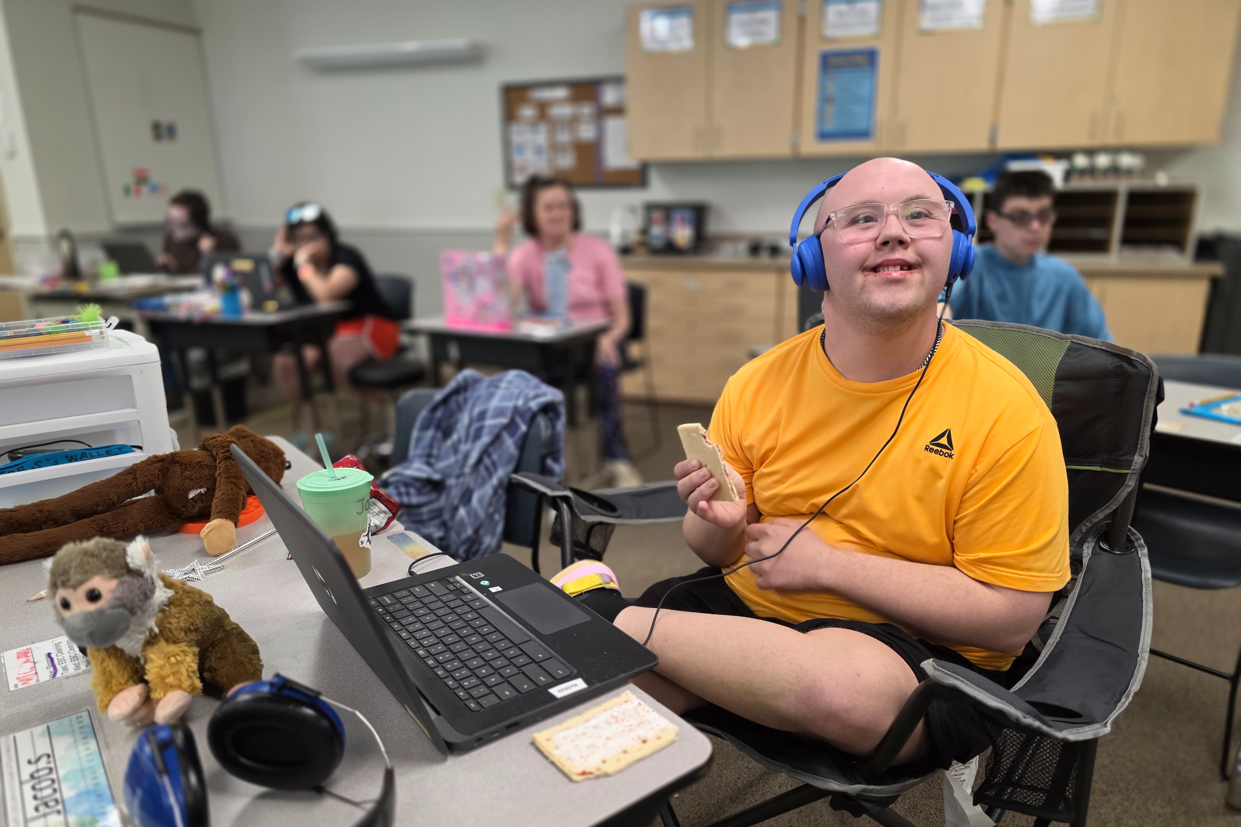 Student sitting at a classroom desk, wearing blue over-ear headphones and glasses, working on a laptop while holding a snack. The desk contains a pair of headphones, a stuffed animal, a green cup, and school supplies. Other students and classroom cabinets are visible in the softly blurred background. Provide your feedback on BizChat