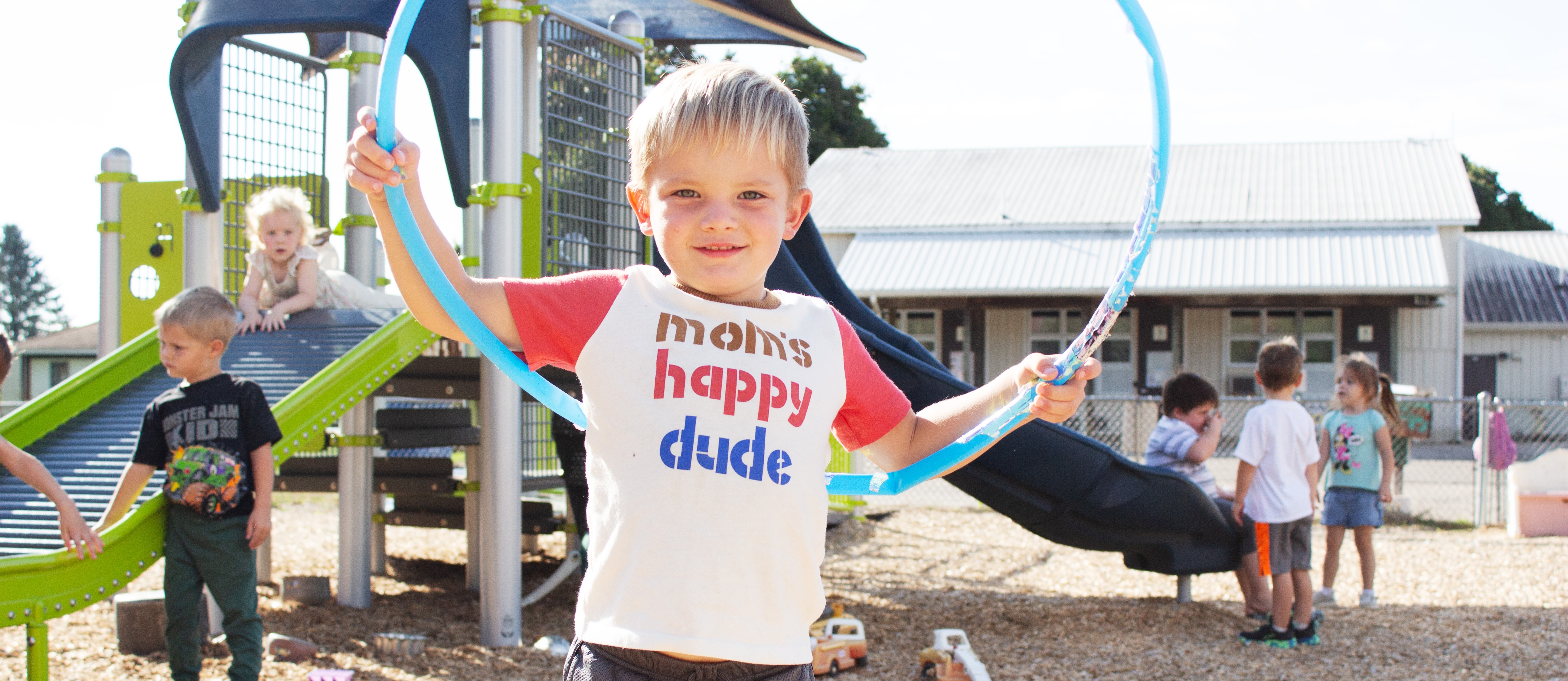 A young boy holds a hula hoop in a playground. Behind him, other children play on slides and equipment.