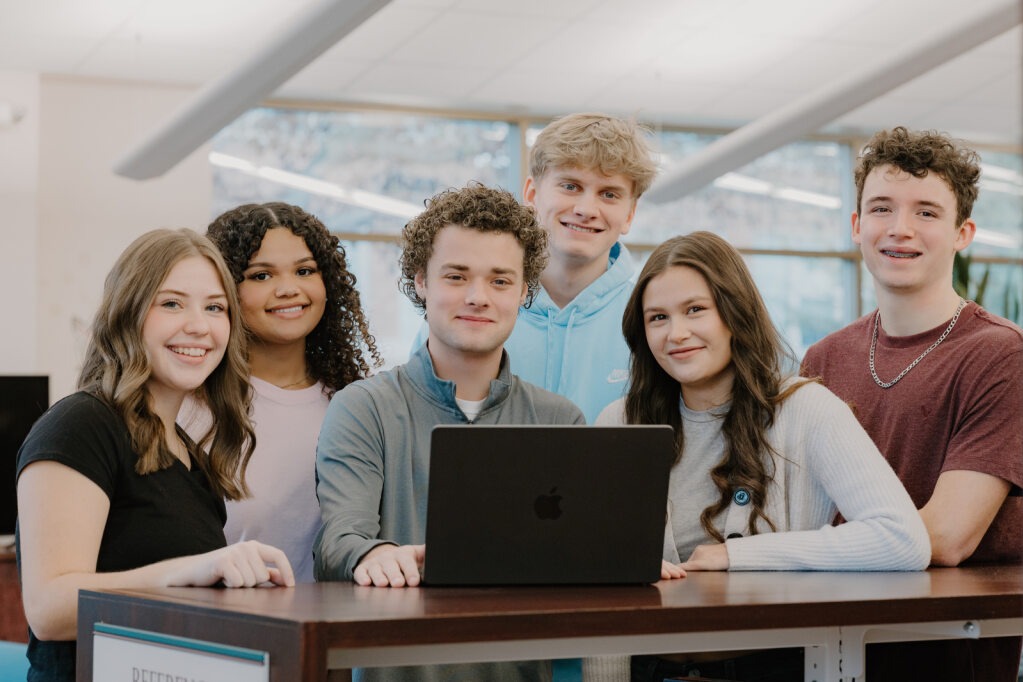 Group of young people with laptops and smiling faces inside an office. The person in front has curly hair.