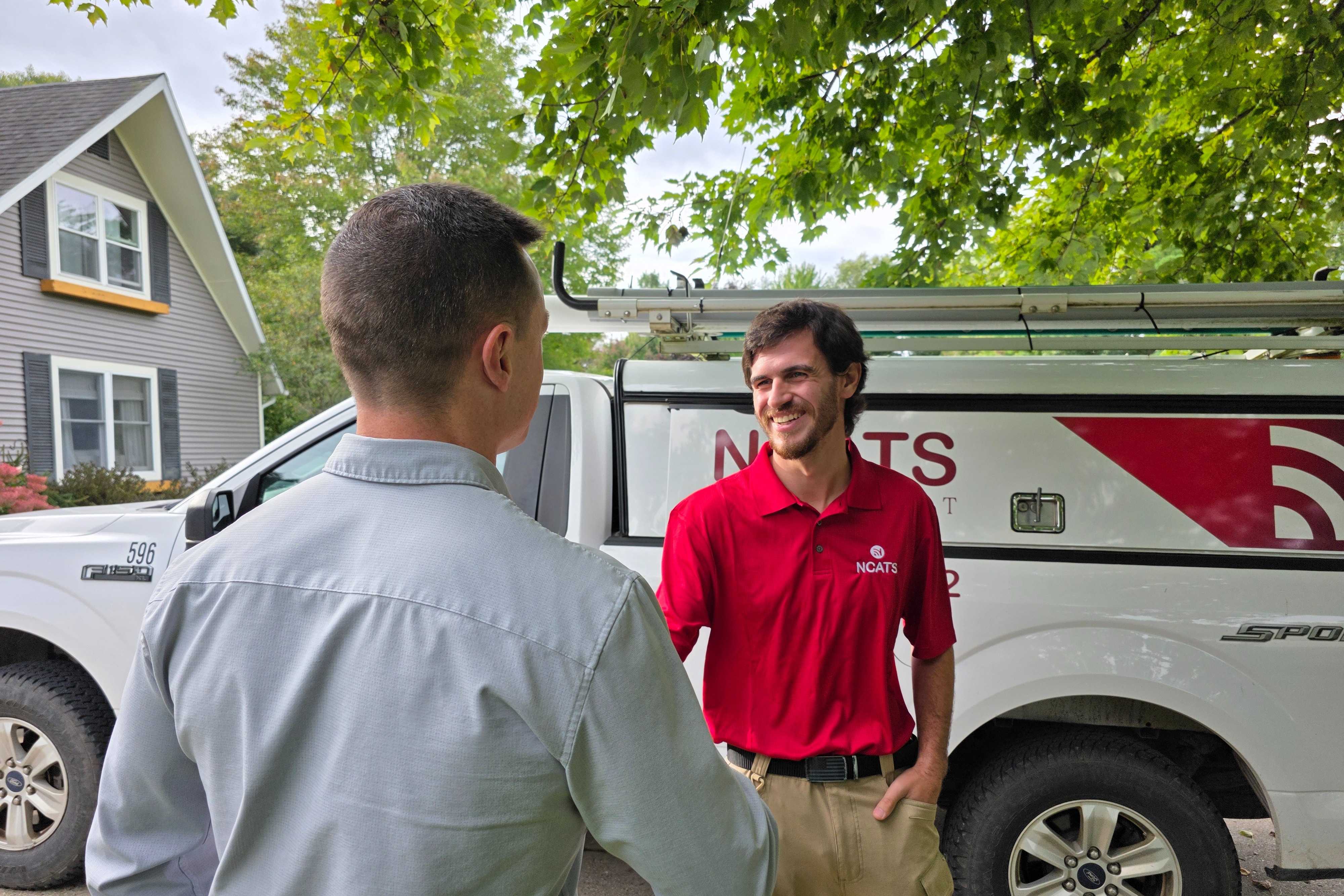 Two men standing in front of a white service vehicle. One wears a red polo shirt and the other a light blue long-sleeve shirt.