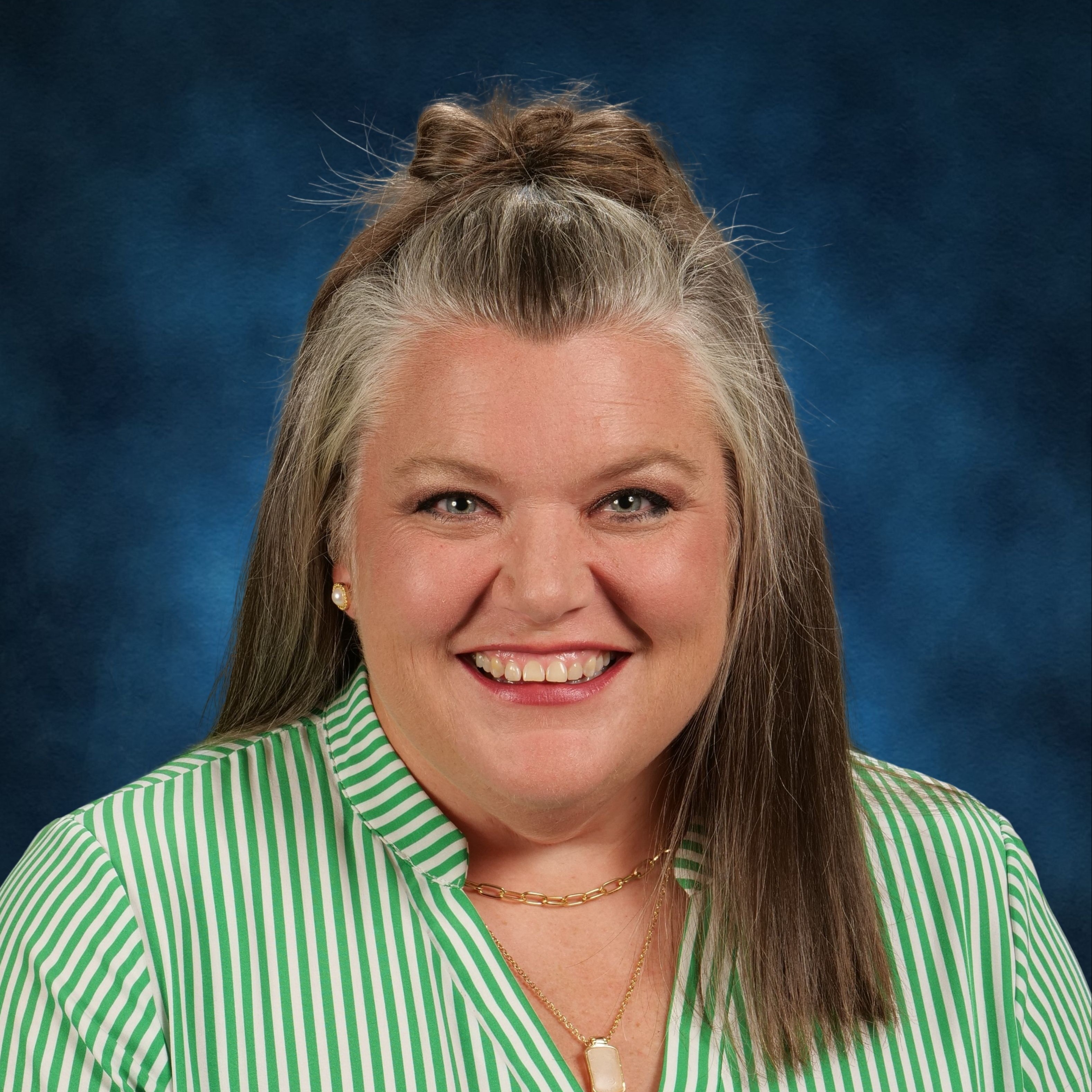 Woman with gray hair in a bun smiles at the camera. She wears a green striped shirt and gold jewelry.