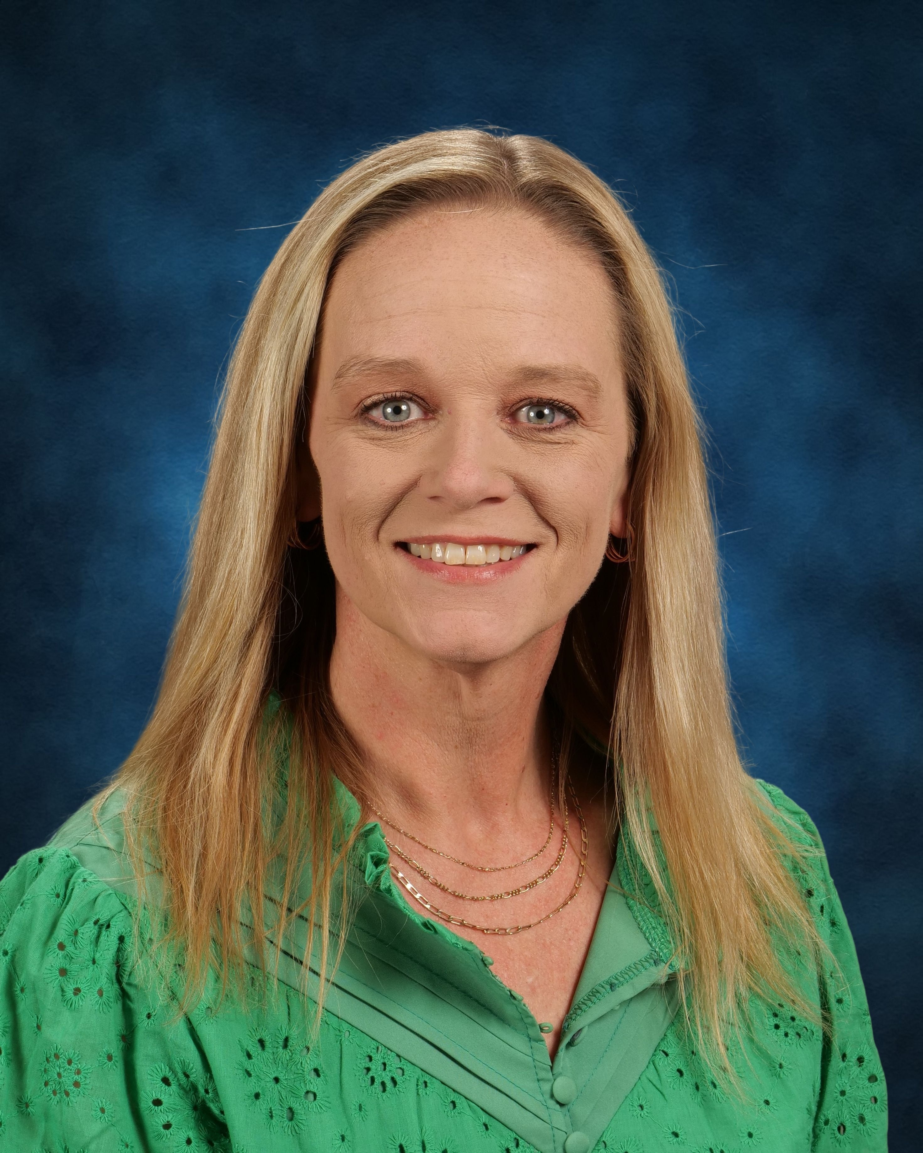 A woman with blonde hair in a green blouse, smiling at the camera. Blue background.