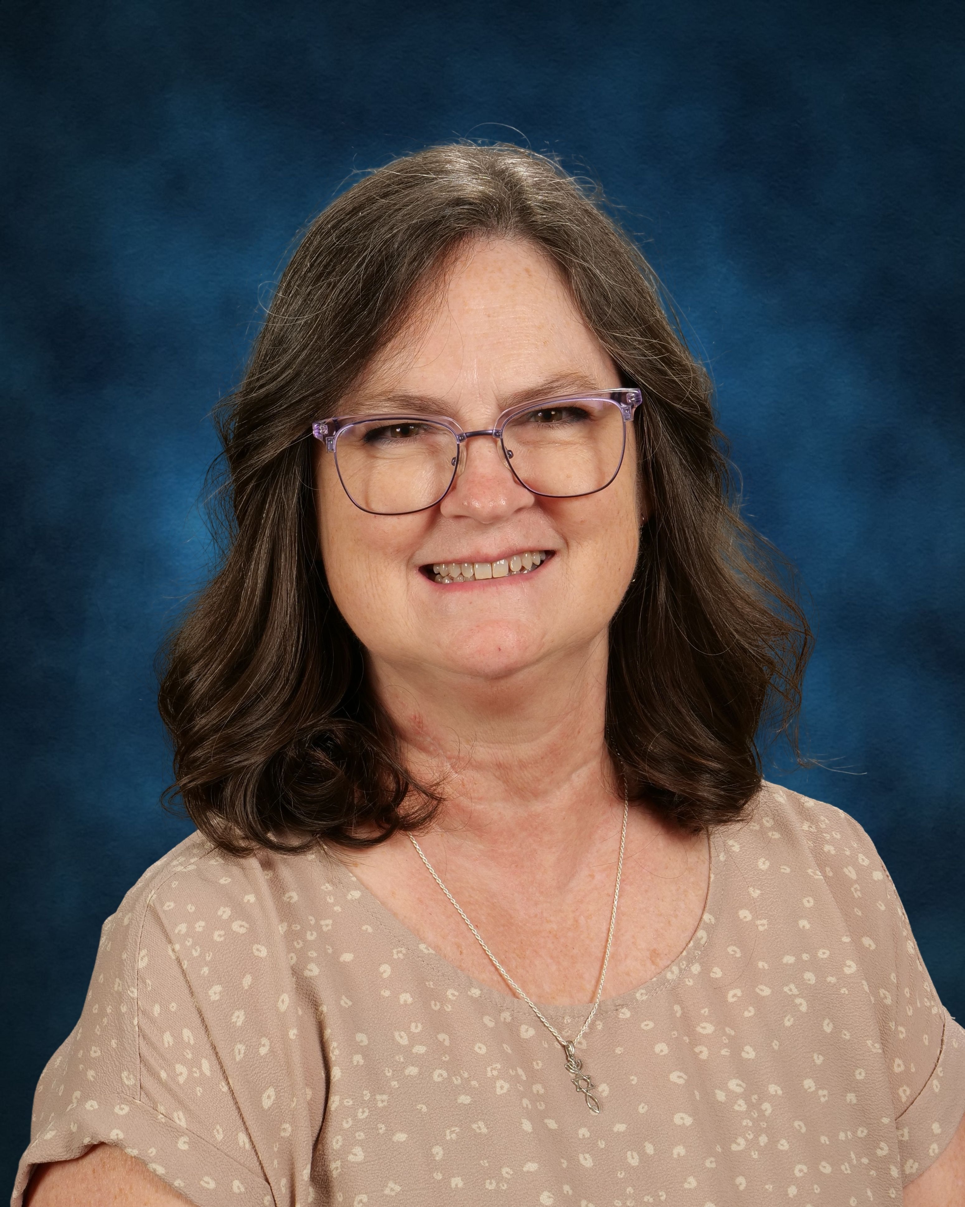 An adult woman with brown wavy hair wearing glasses, a beige top with small white dots, and a necklace.