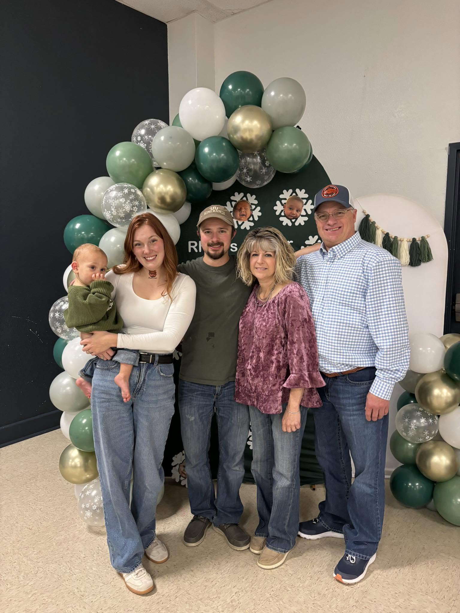 Five people stand in front of a decorative balloon arch. A woman holds a baby. Two men wear hats. Two women wear blouses.
