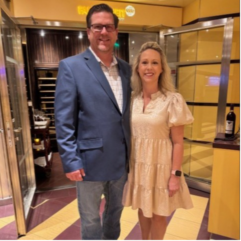 A man and a woman stand side by side, smiling. The man wears a blue suit and glasses. The woman has blonde hair and a white dress. Behind them is a room with glass walls.
