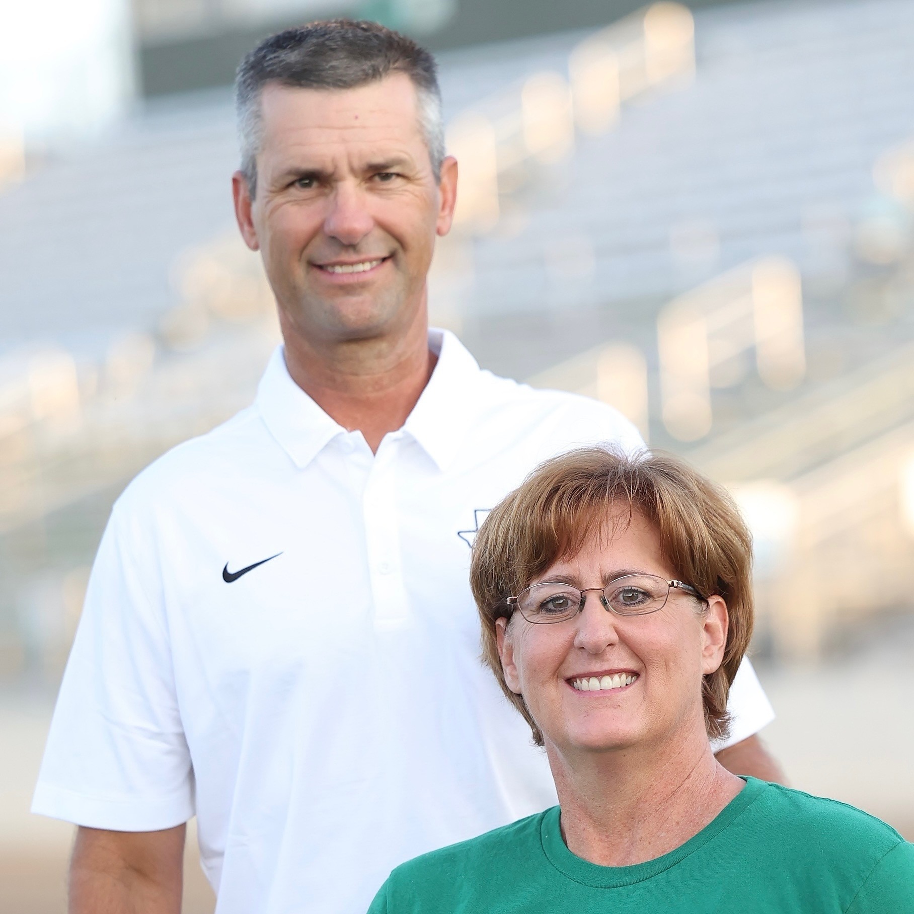 A man and a woman smile for the camera. The man wears a white polo, the woman wears a green T-shirt. Behind them are empty stadium bleachers.