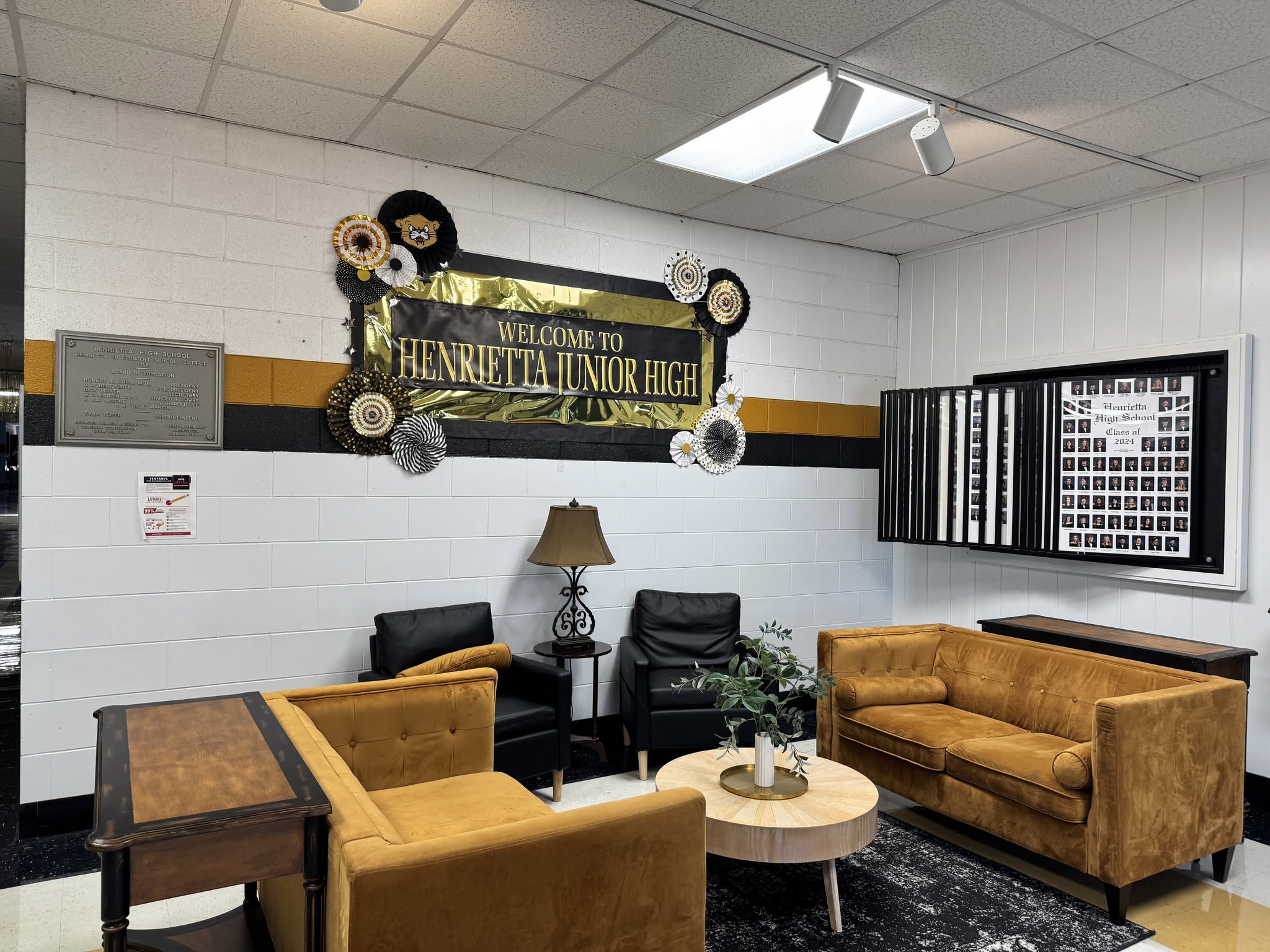 A room with furniture including couches, a table, and a lamp. A sign on the wall reads "Welcome to Kennetha Public High."