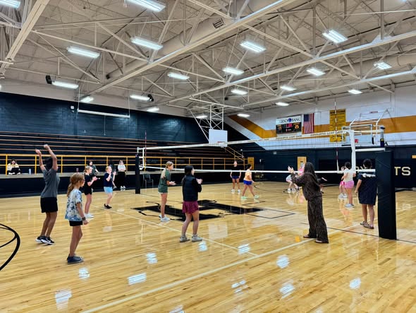 An indoor court with multiple people playing volleyball. Bright lights on the ceiling. Blue walls and wooden floor.