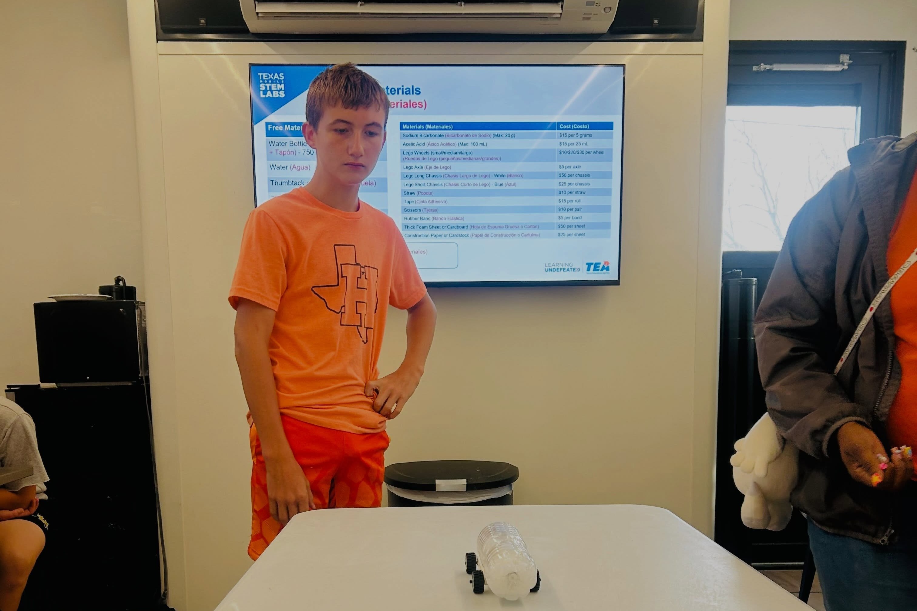 A person stands in front of a table with a monitor behind them. An individual is nearby, holding an object.