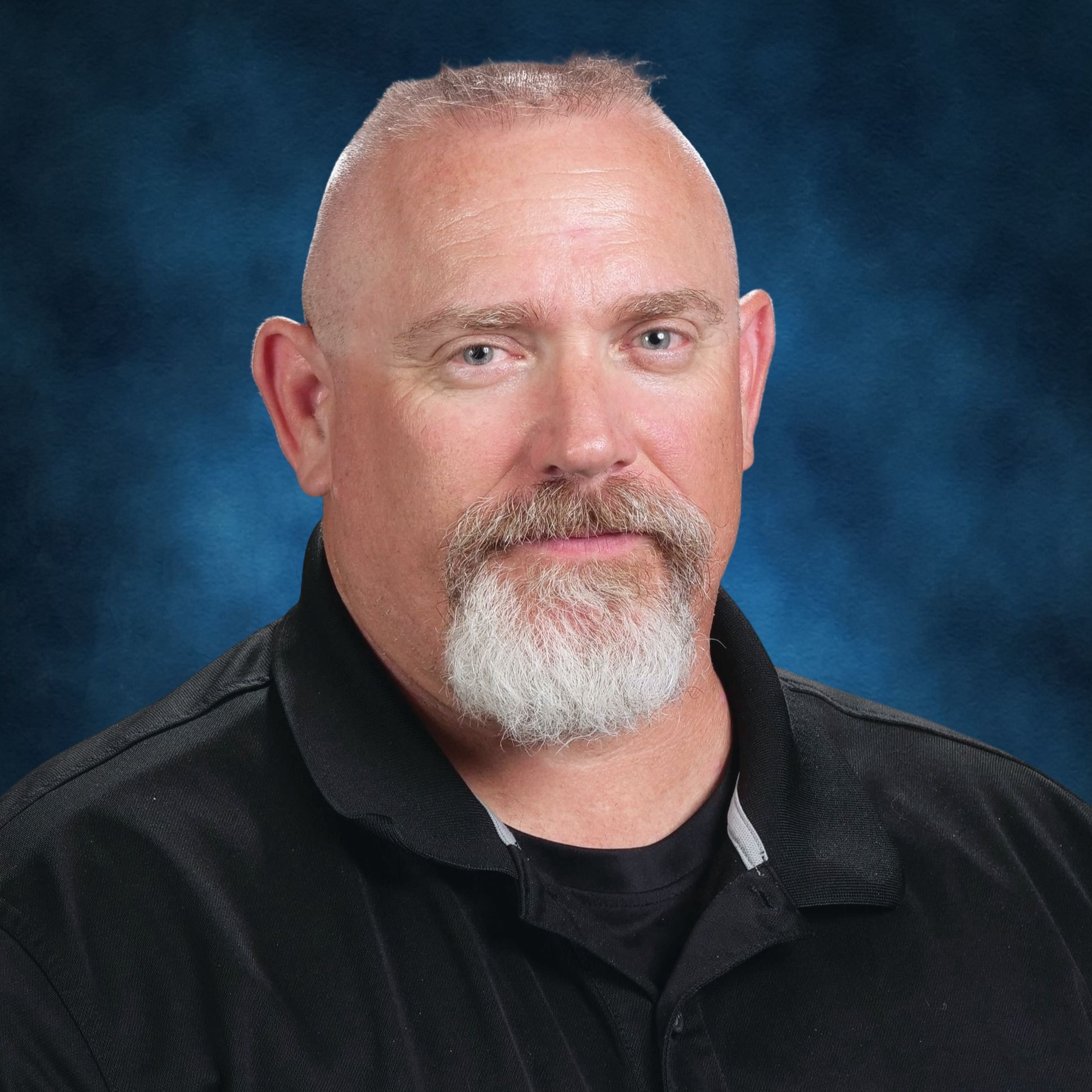 A man with a white beard and short gray hair, wearing a black polo, looks directly at the camera.