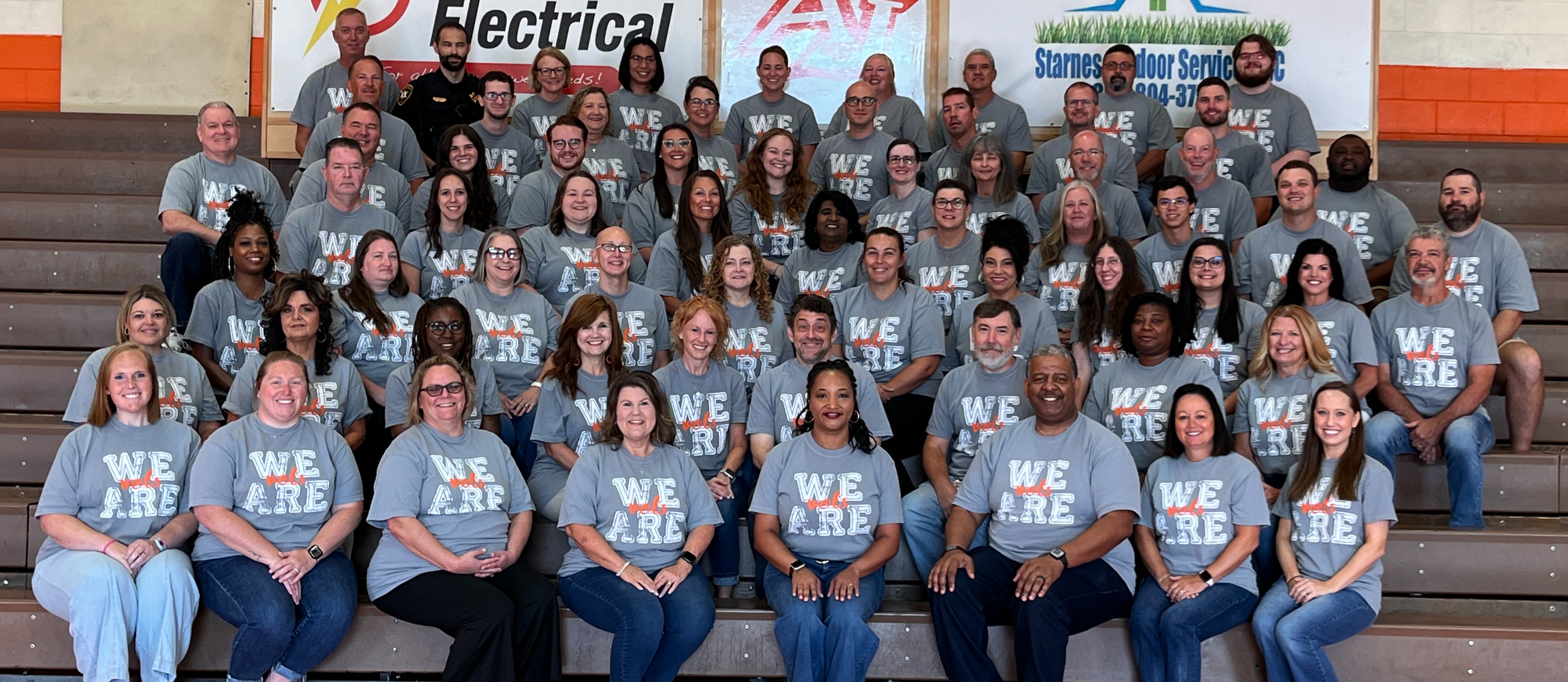 A group of adults in matching shirts with "WE ARE" written on them, posing for a photo in a gymnasium.