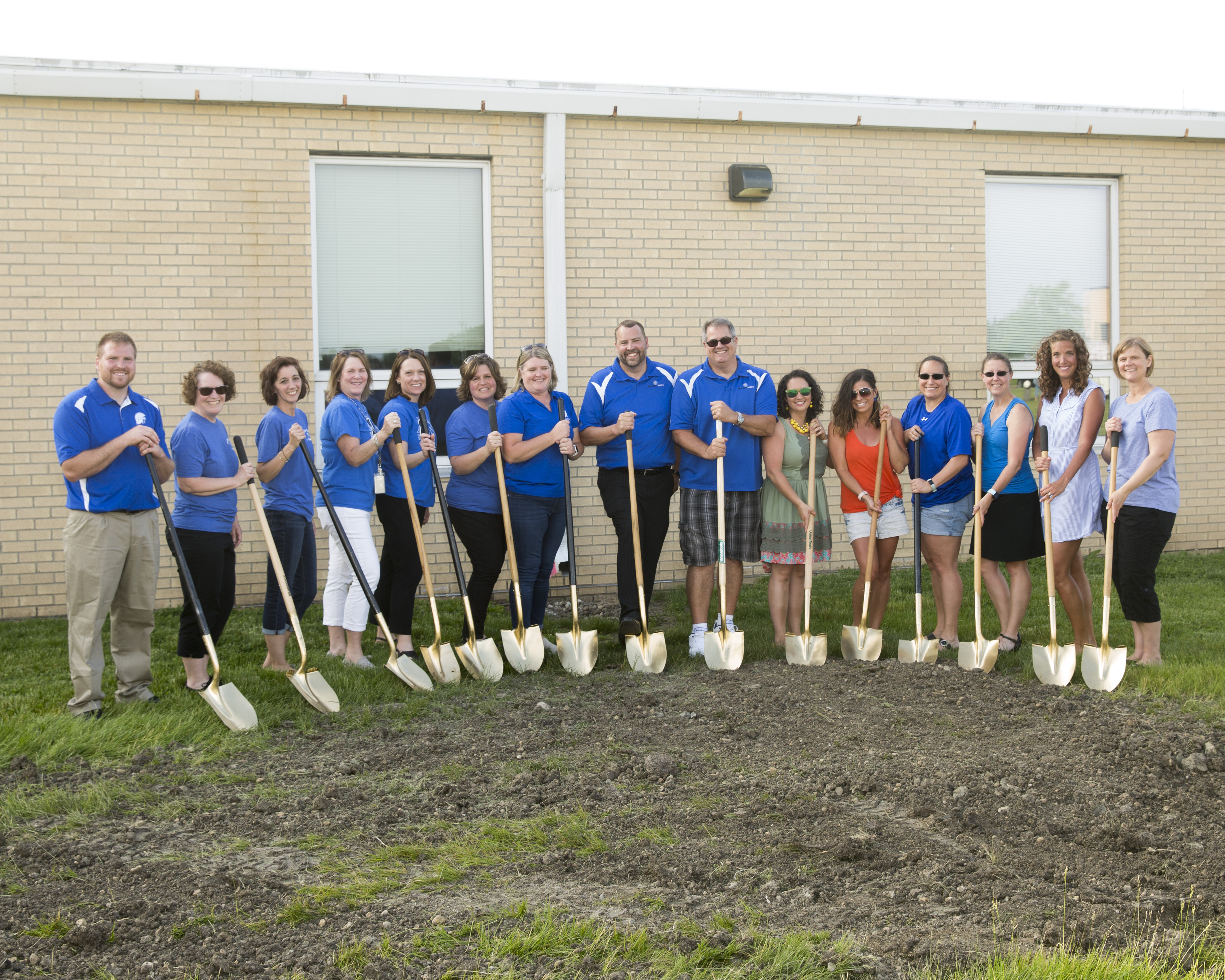 A larger group of teachers with the same blue polo shirt and holding up a set of shovels as well.