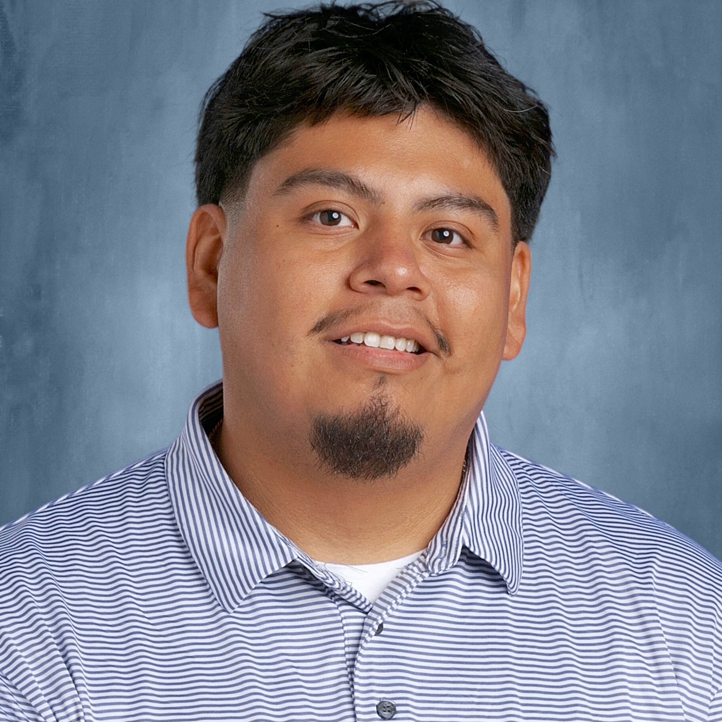 A man with a beard and mustache smiles at the camera. He wears a striped shirt with a logo.
