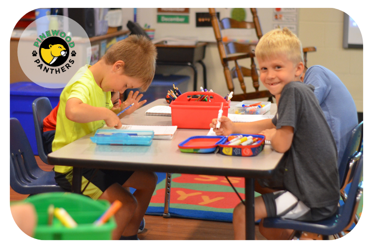 pinewood students coloring at a classroom table and one student smiling at the camera.