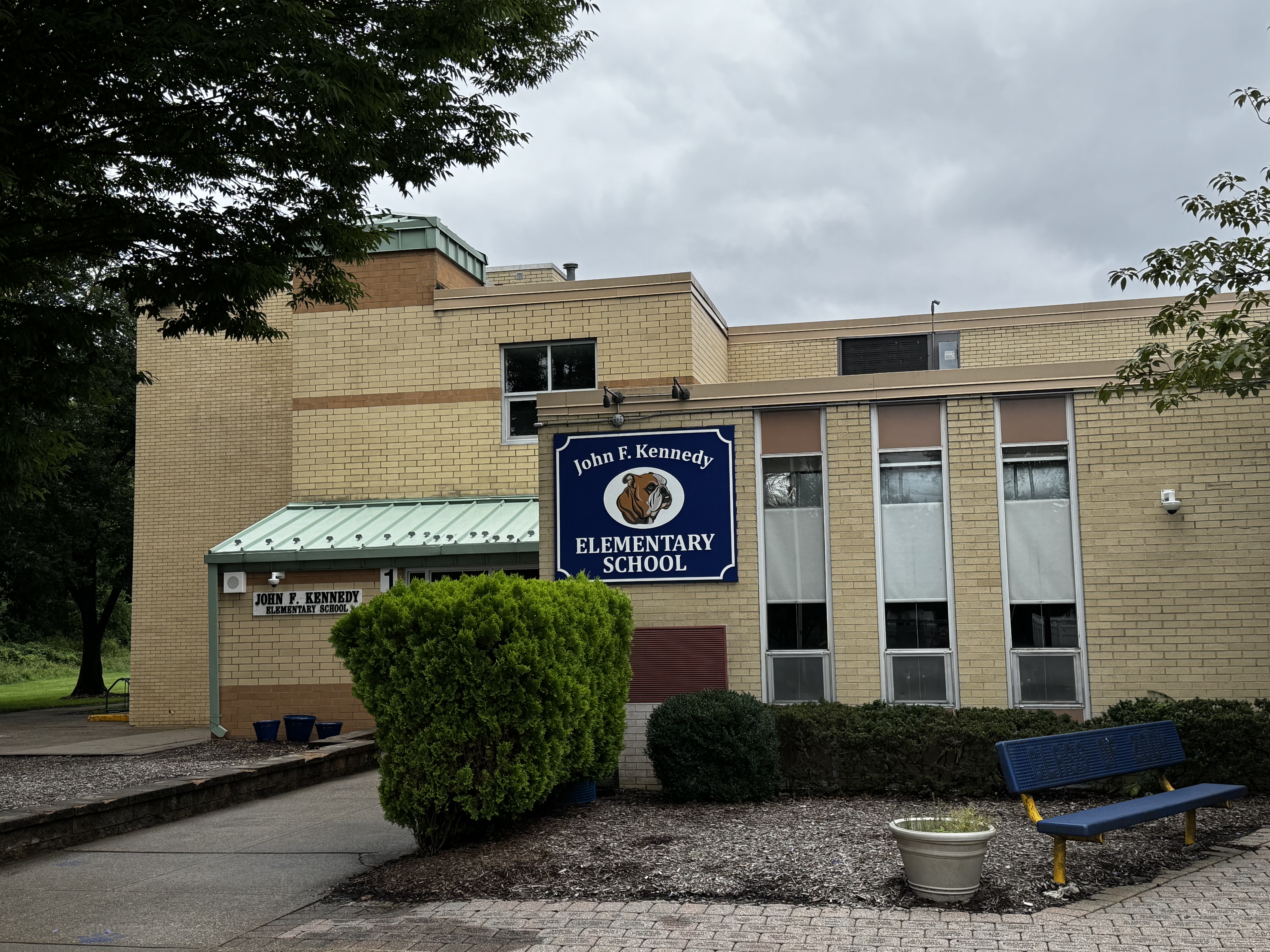 A blue sign reading John F. Kennedy Elementary School above a brick building with several windows. A bench is in front of the building.
