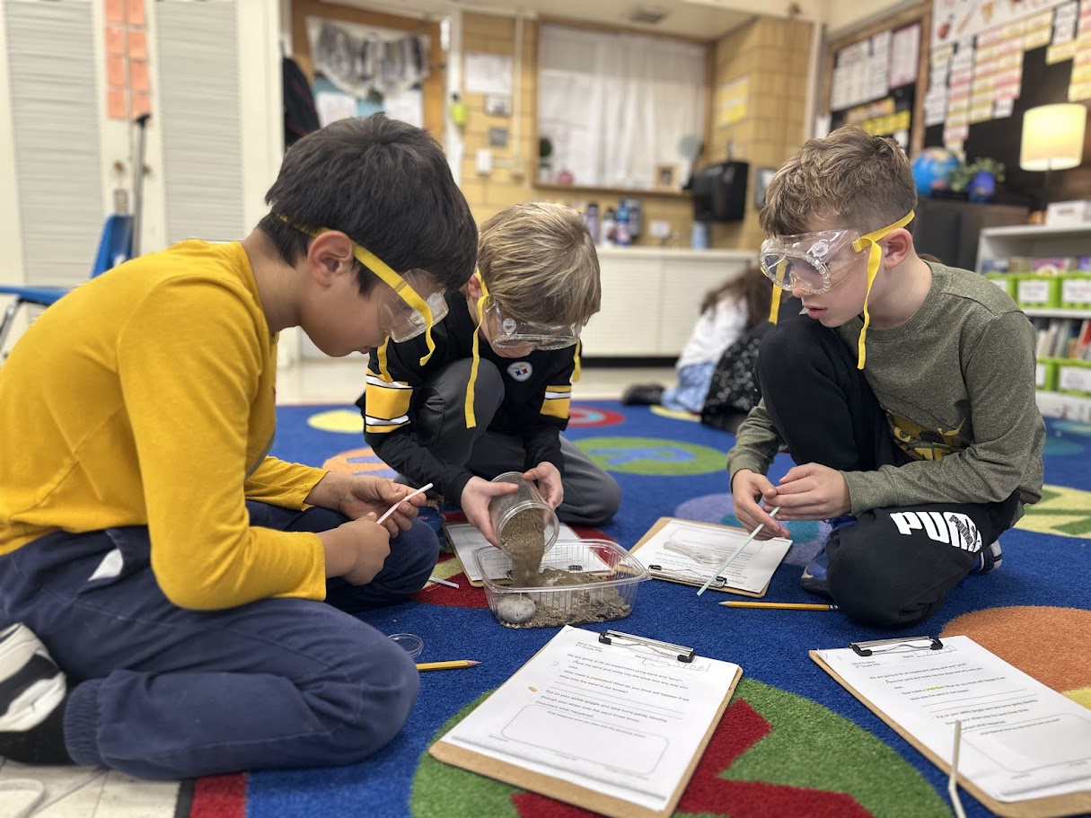 Four boys wearing goggles, sitting on a colorful carpet, doing an experiment with a bowl and papers.