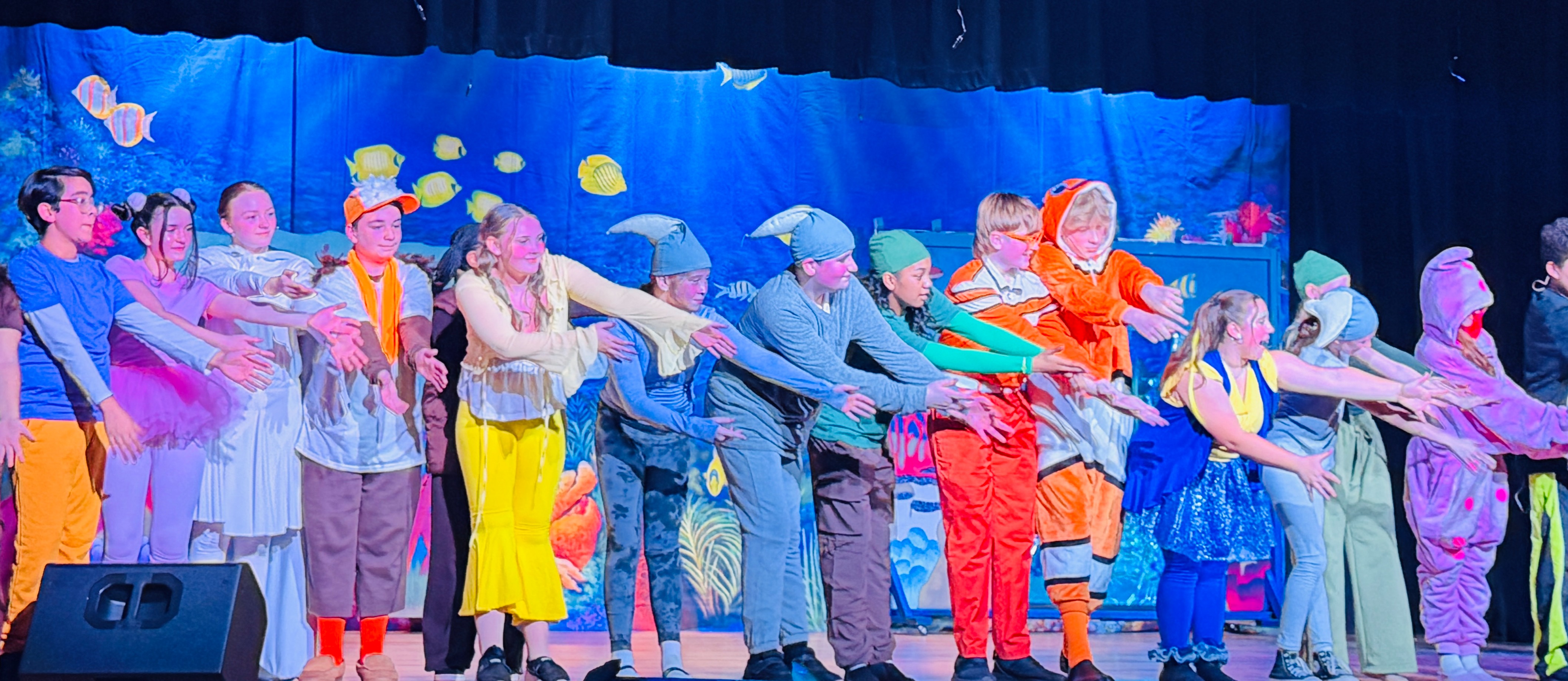 A group of people dressed in colorful costumes performs on stage, with an audience watching.