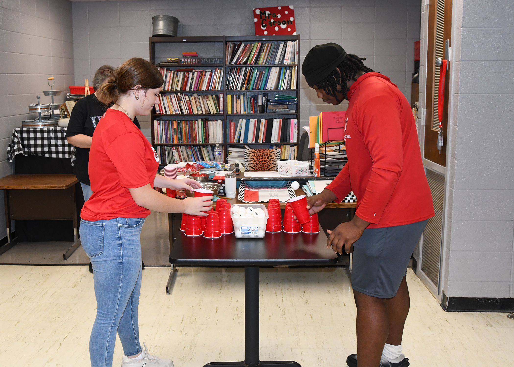 Two individuals in red shirts stack red cups on a table in a room with bookshelves.