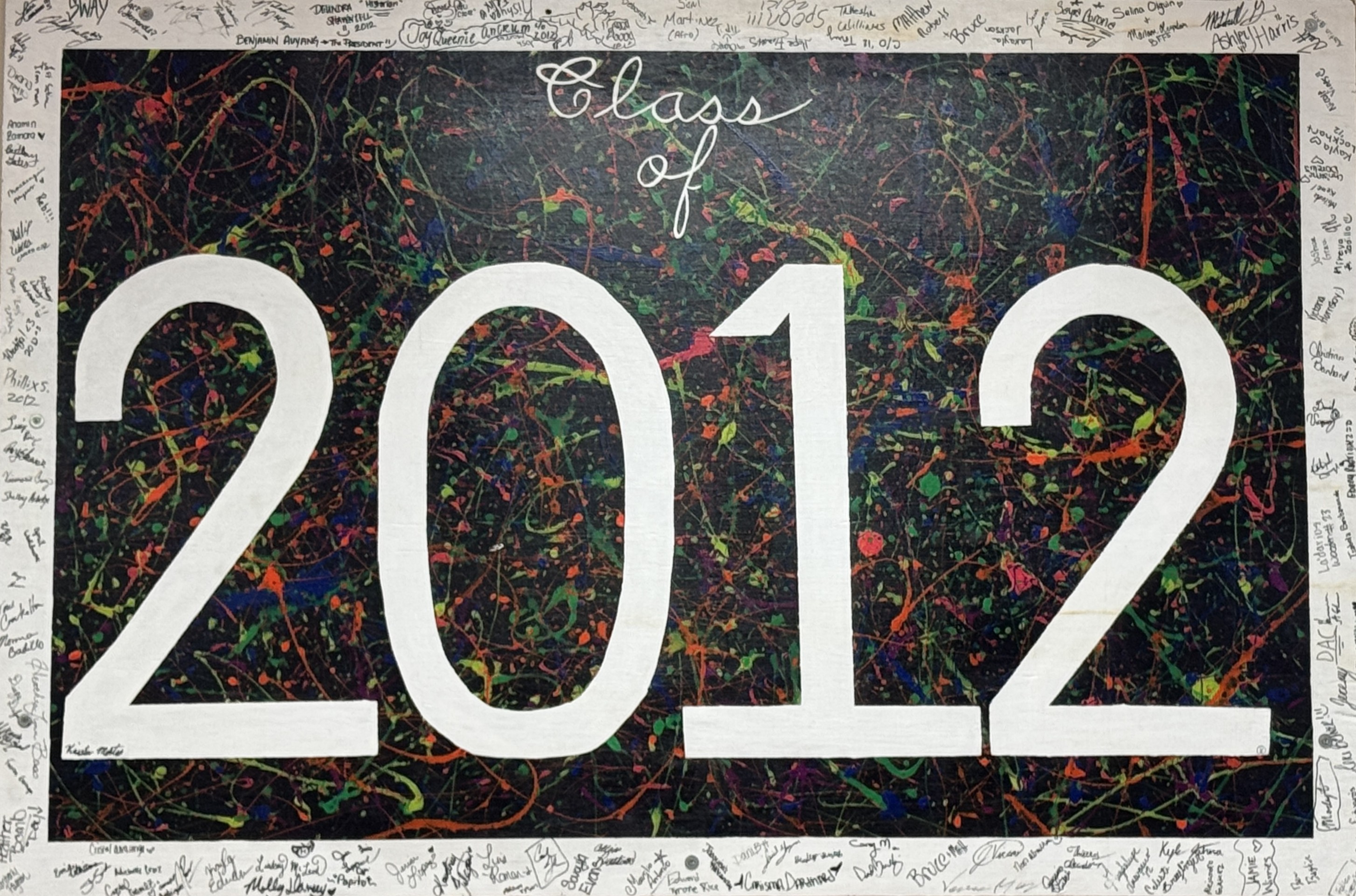 A colorful poster featuring the text 'Class of 2012' in large white numbers and letters against a black background splattered with vibrant paint colors like green, orange, purple, and blue. The border around the poster is white and filled with handwritten signatures and small doodles from students.