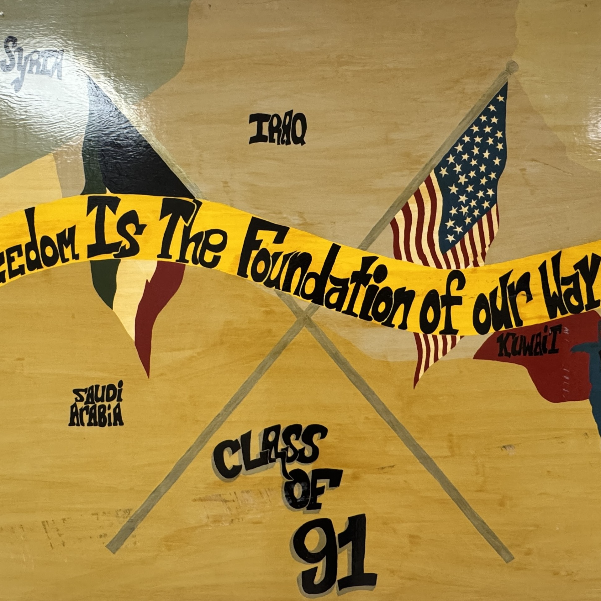 A mural-style painting of a Middle East map with labeled countries including Iraq, Iran, Kuwait, Saudi Arabia, Jordan, Israel, Lebanon, Syria, and others. The Persian Gulf and Red Sea are also labeled. Two crossed flags are featured—one of the United States and one with red, white, black, and green stripes, representing a Pan-Arab flag. A yellow ribbon across the center reads, "Freedom Is The Foundation of our Way of Life." At the bottom, it says "Class of 91" in bold black letters.