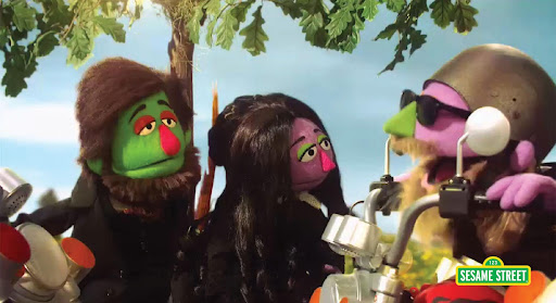 Three colorful puppets interact under a tree, with one wearing a helmet, while another holds a guitar and the third has an expressive demeanor.