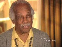 An elderly person wearing a yellow shirt and a gray cardigan, seated and appearing in a warmly lit indoor setting.