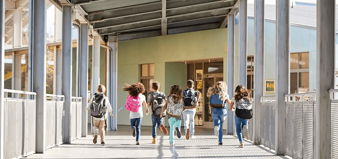 A group of children run in a hallway with metal railings and buildings in the background.