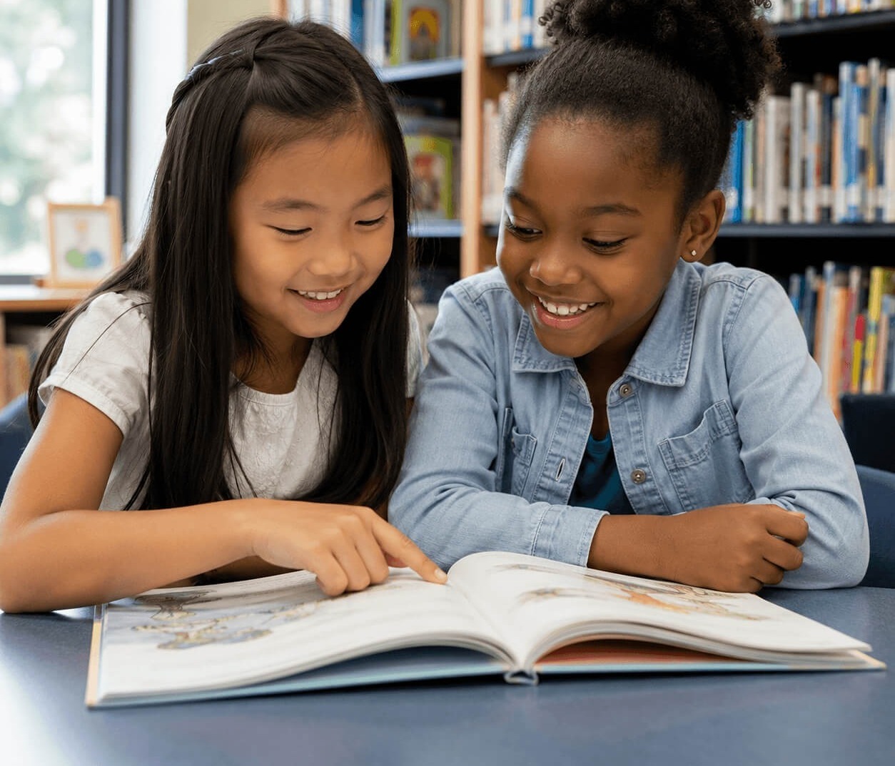Two girls seated at a table, smiling while reading a book in a library with bookshelves.