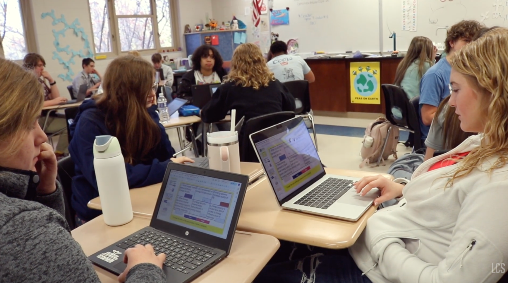 A classroom with students seated at desks. Four people are using laptops. A whiteboard is visible in the background.