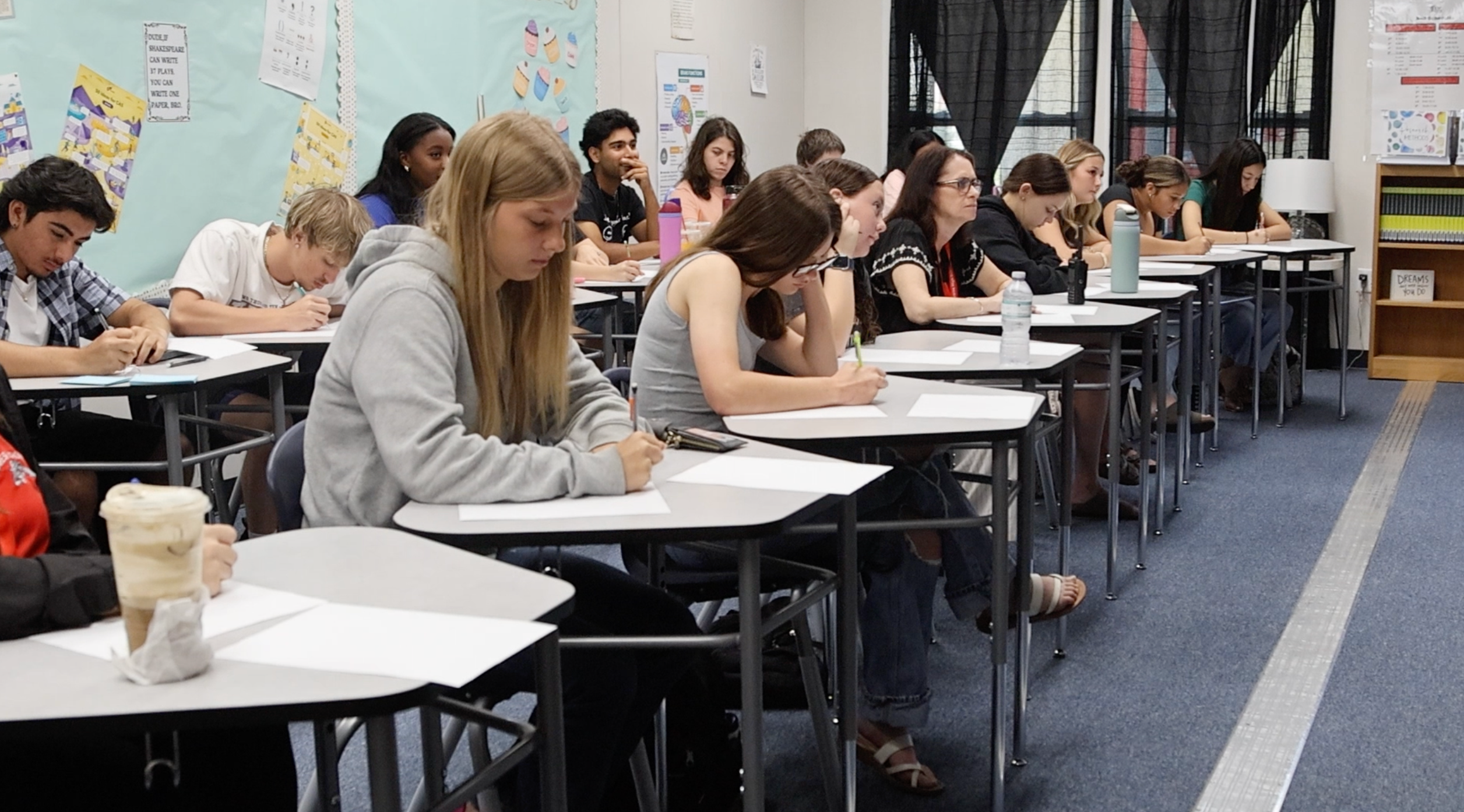 Students sit at desks, writing in notebooks. One student drinks coffee. Desks are arranged in rows.