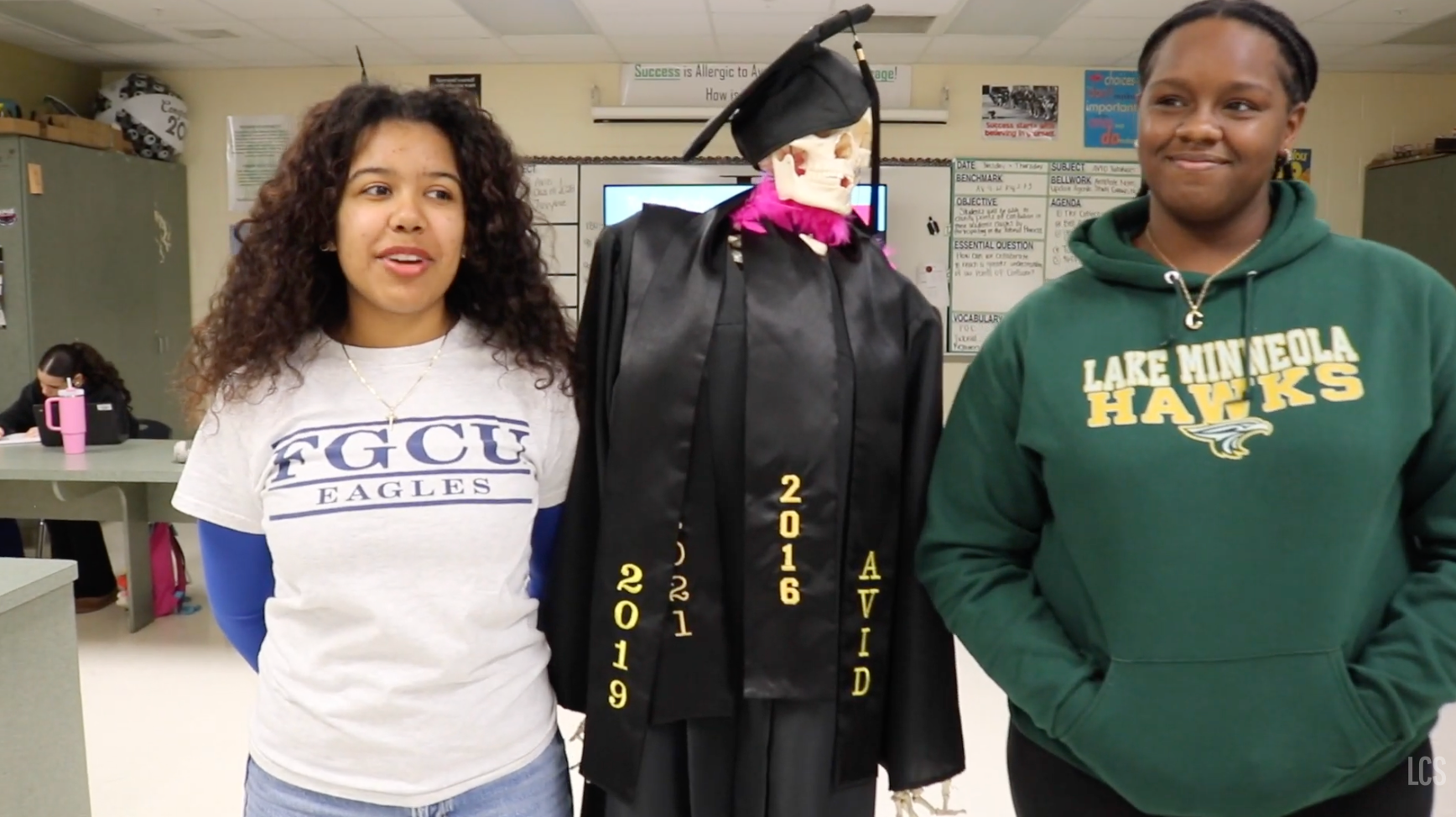 Three women stand next to each other; one wears a cap and gown, another a green hoodie, and the third a white shirt.
