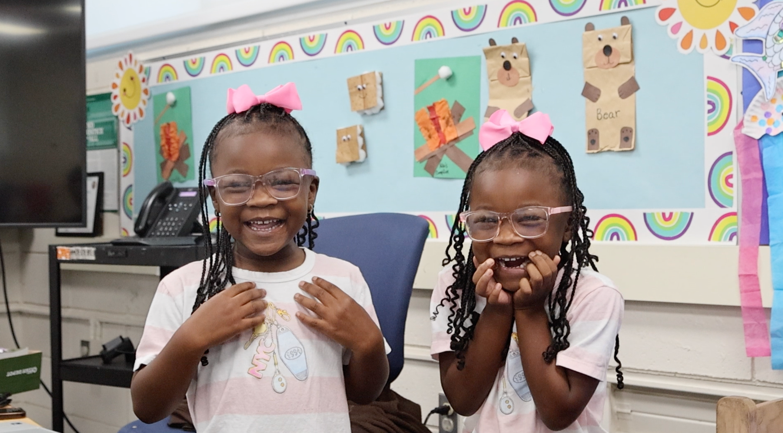 Two young girls with glasses, one with pink bows, stand next to each other and smile.