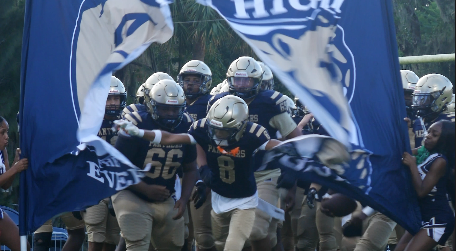 Football players wearing helmets and jerseys run onto the field. Two large flags are held in front of them.