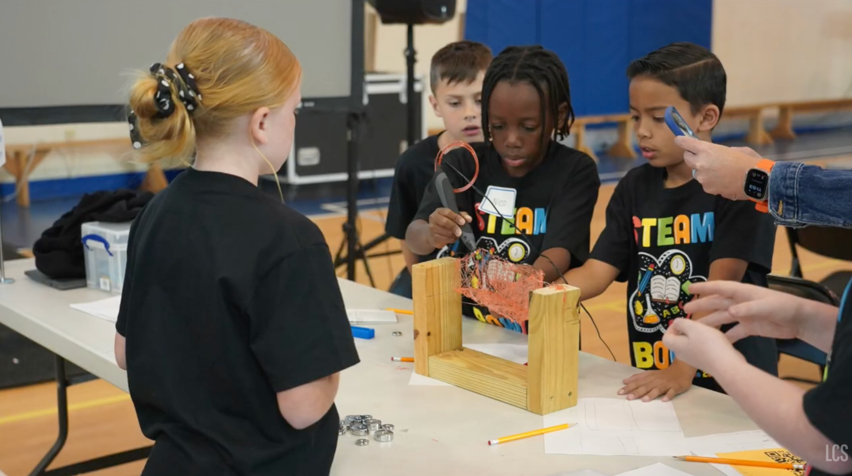 Four kids in black shirts, wearing watches, work on a project at a table. One child holds a tool.