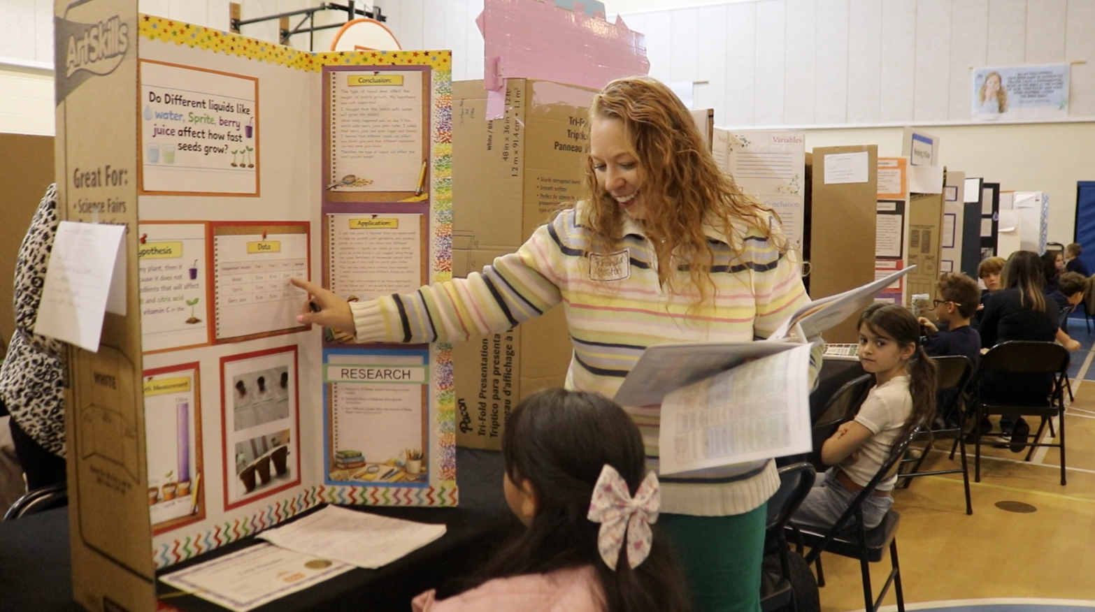 A woman stands by a cardboard display board, pointing at it while holding a folder. Children sit in chairs behind her.