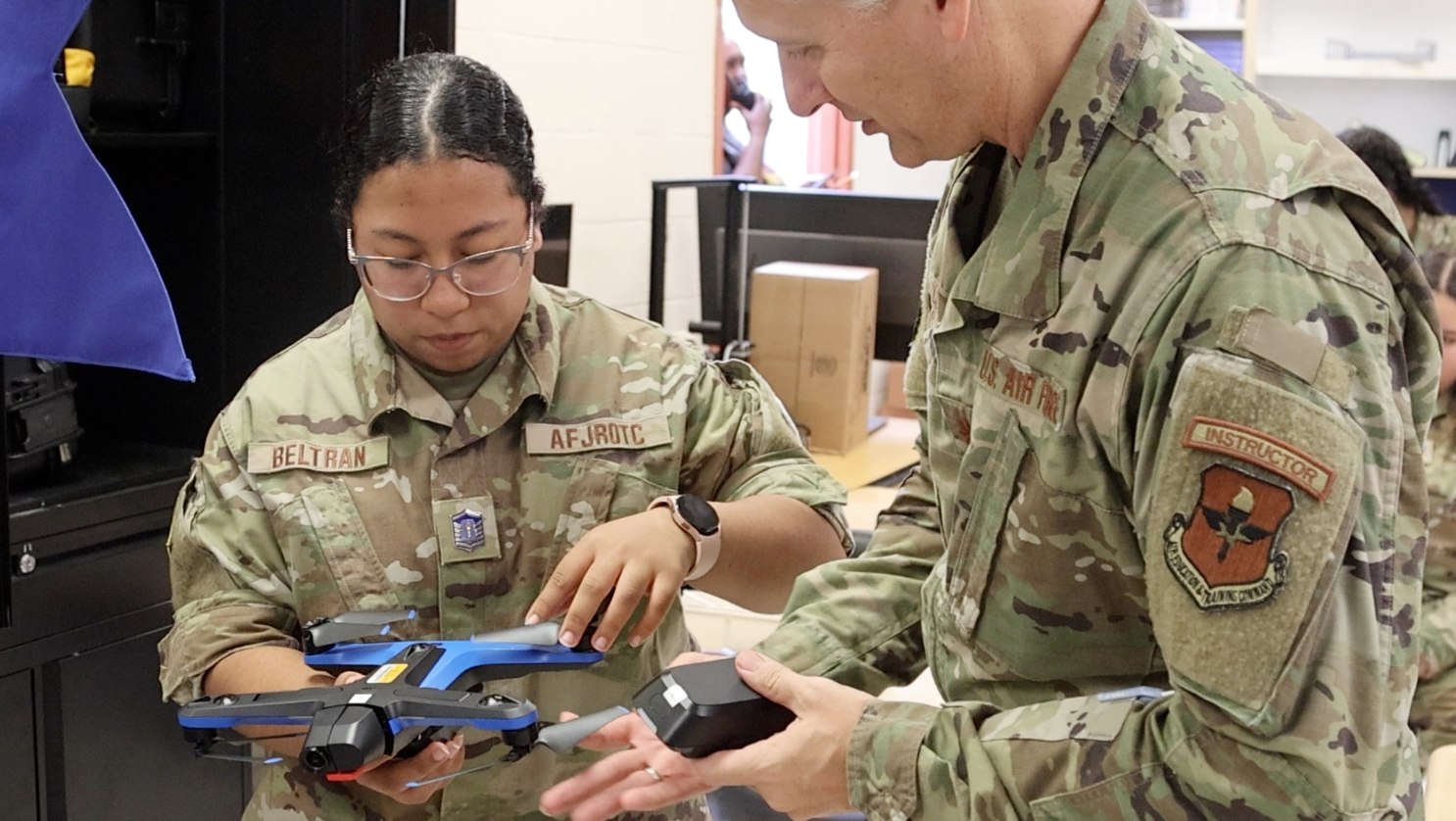 Two military personnel in uniform, one holding a blue drone and the other a black device, with a wooden box behind them.