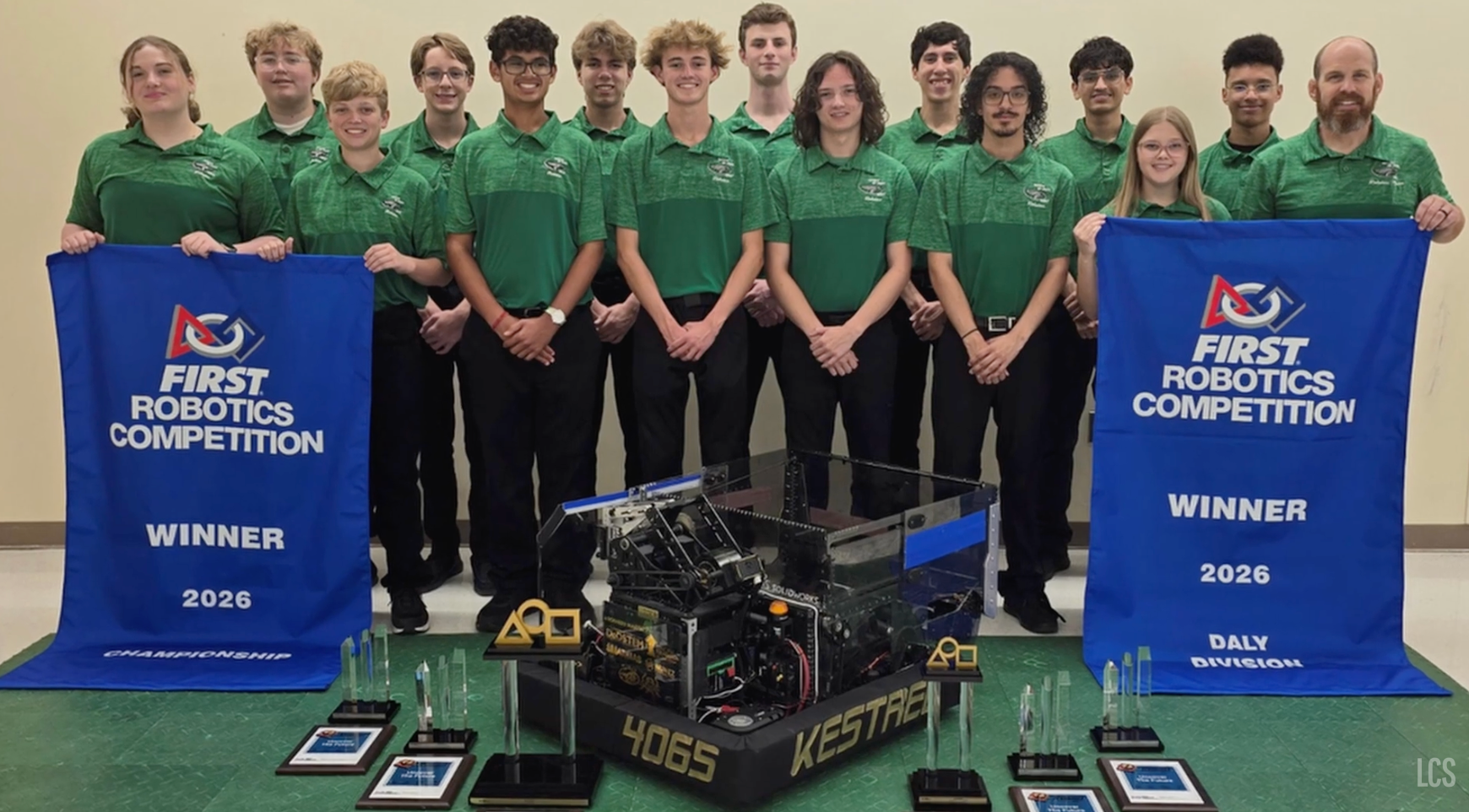 A robotics team in green uniforms poses with a robot and banners. One banner reads "FIRST Robotics Competition" and "WINNER 2016".