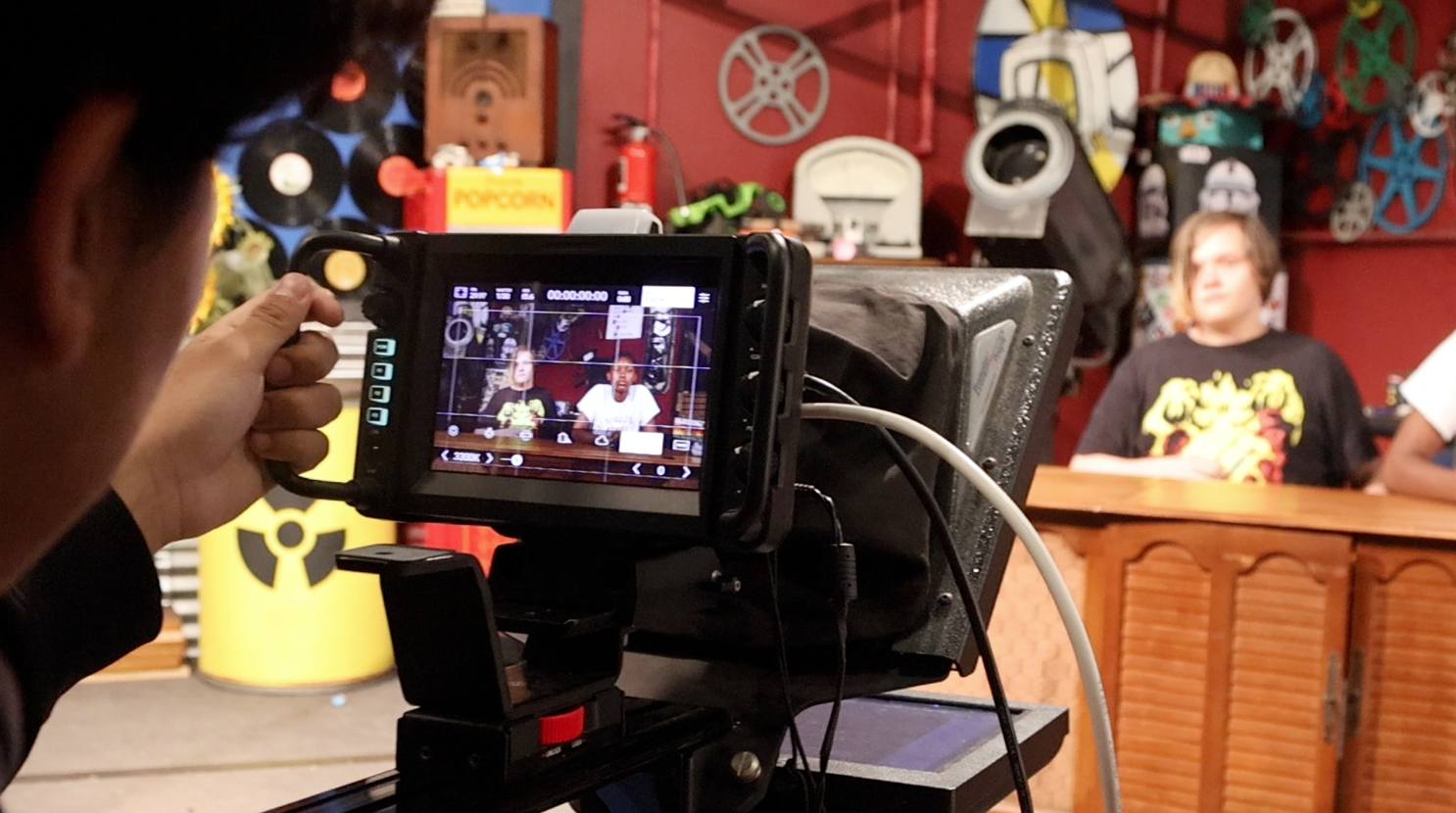 A person holds a camera filming two people seated at a desk. The desk is brown.