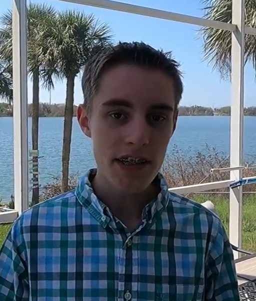 Man in a plaid shirt stands in front of a lake with trees and blue sky.
