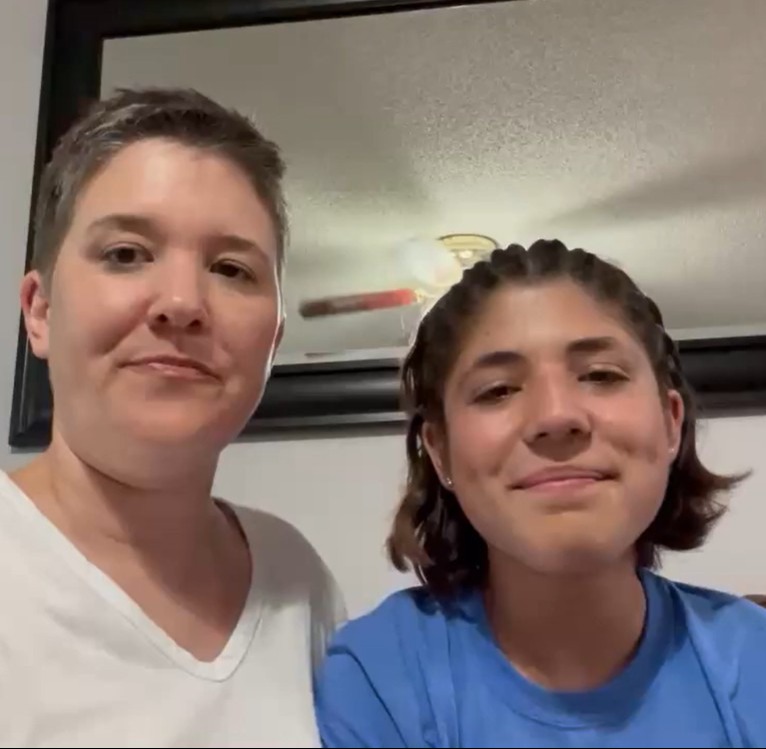 Two women stand close, facing camera. One wears a white shirt; the other wears a blue shirt. A picture frame and a ceiling fan are in the background.