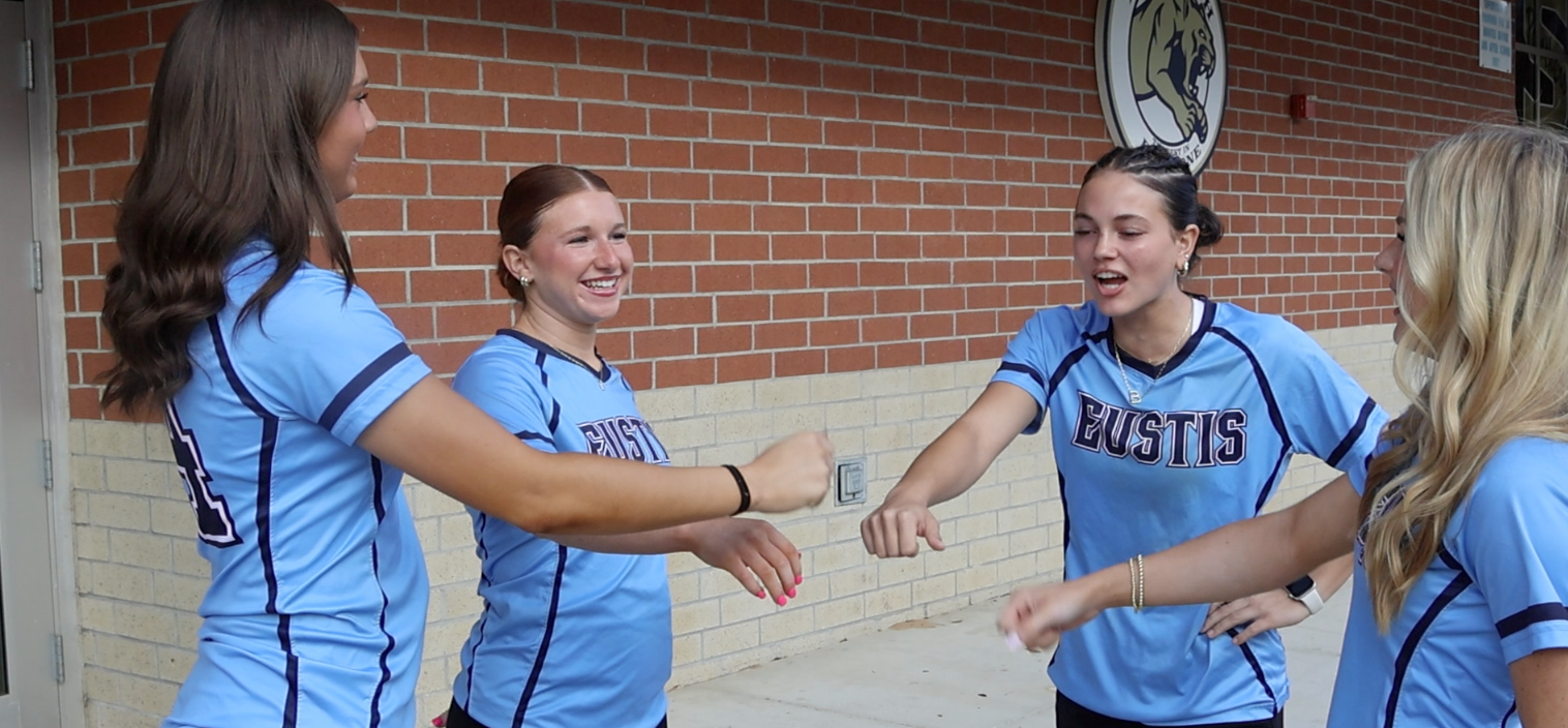 Four women in matching blue jerseys stand together in front of a brick building, sharing a laugh.