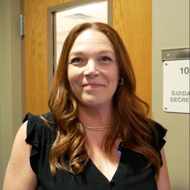 A woman with curly brown hair in a black blouse stands next to a door with a room number sign.