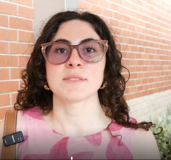 A woman with curly hair wearing glasses stands against a brick wall, holding a black object. She wears a pink floral-print top.
