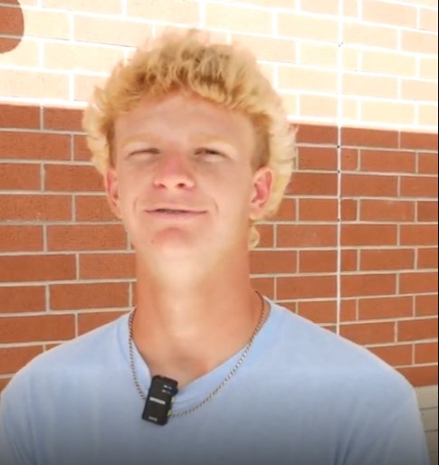 A person with blond hair and a blue shirt stands in front of a brick wall.