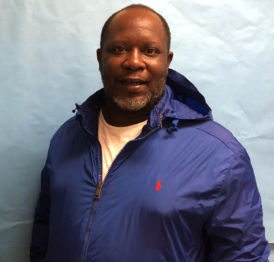 Man in blue jacket with white undershirt, light blue background. He has a beard and a slight smile.