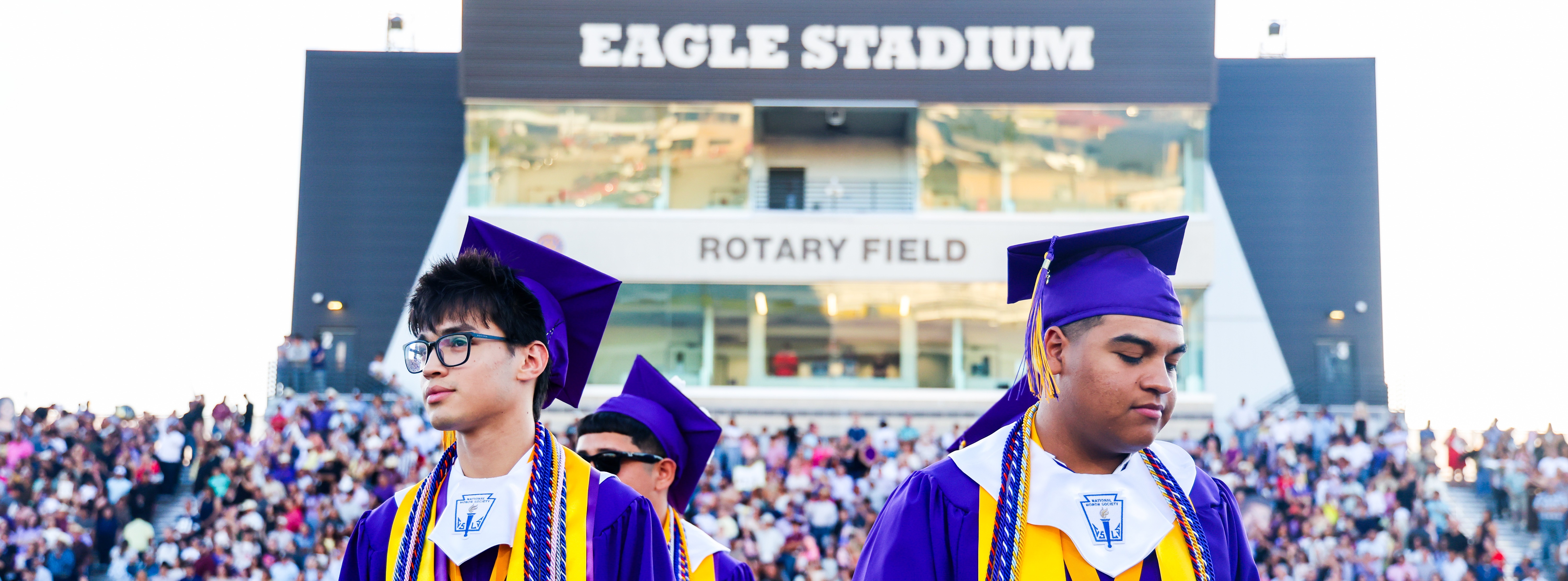 two graduates walk the field with the stadium and crowd in the background of the photo