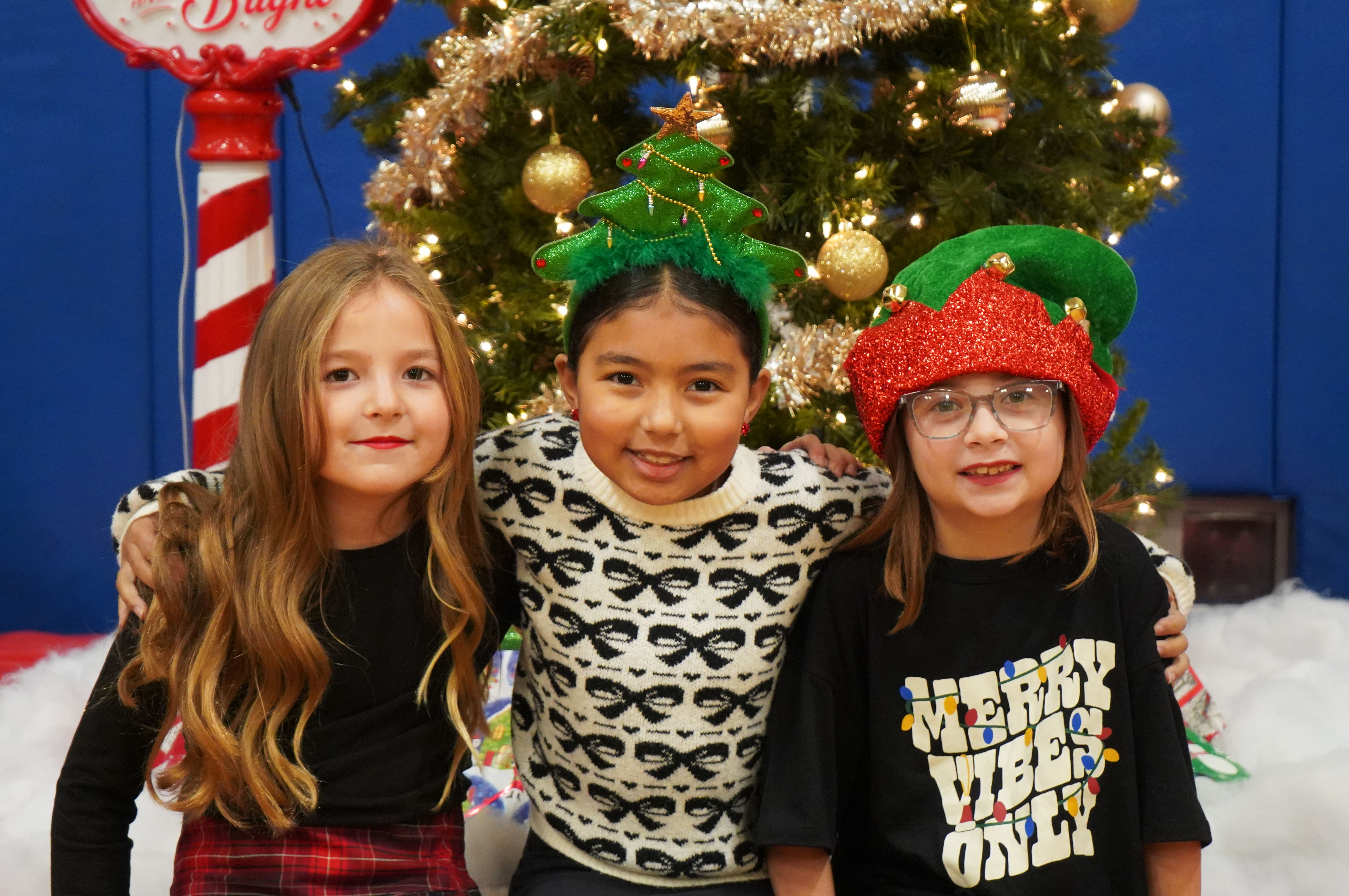 Three third grade friends sit in front of a Christmas tree at Grandparents Breakfast. 