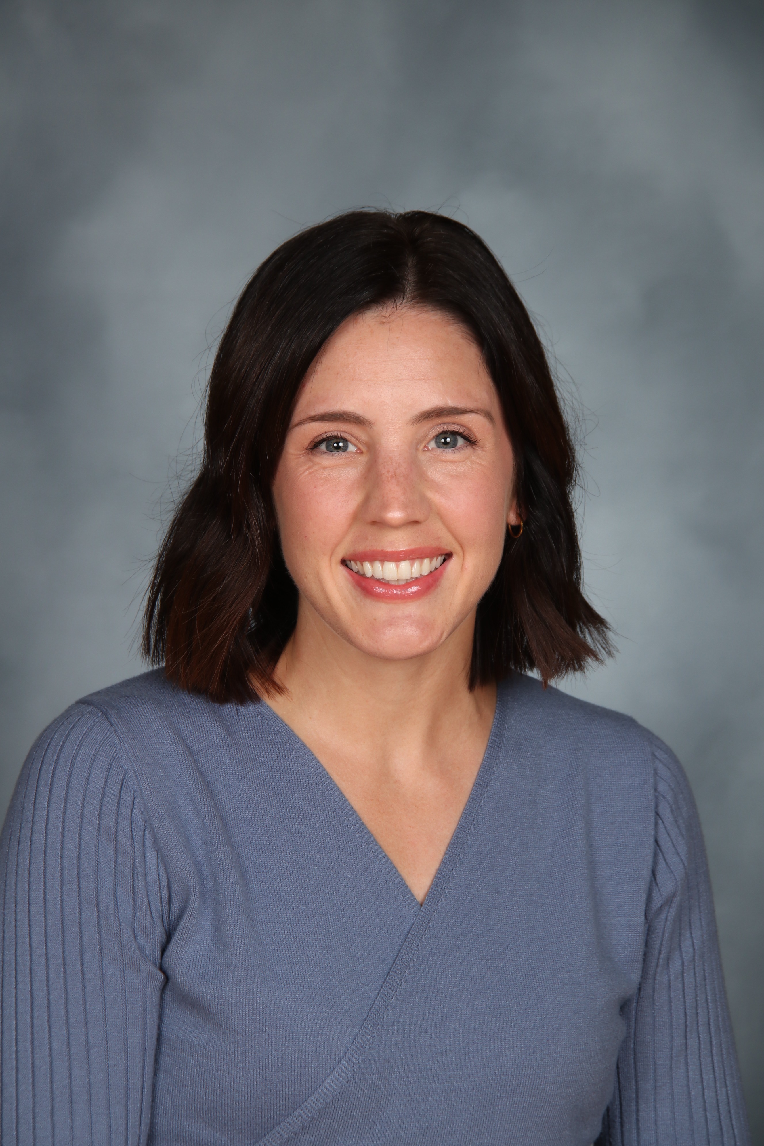 A smiling woman with short dark hair wears a light blue V-neck blouse. Her head and shoulders fill the frame.