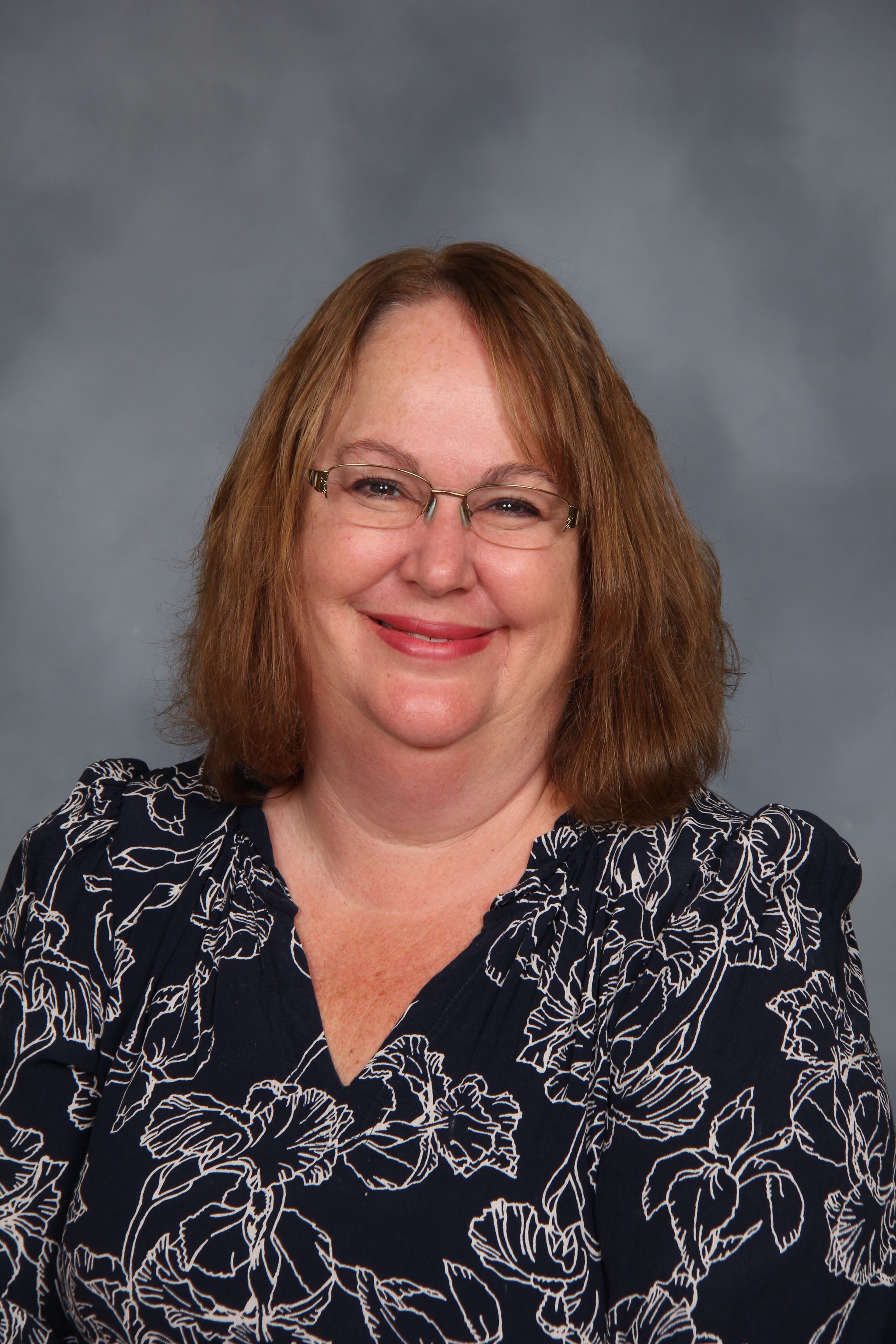 Woman wearing glasses and a floral blouse smiles at the camera against a gray background.